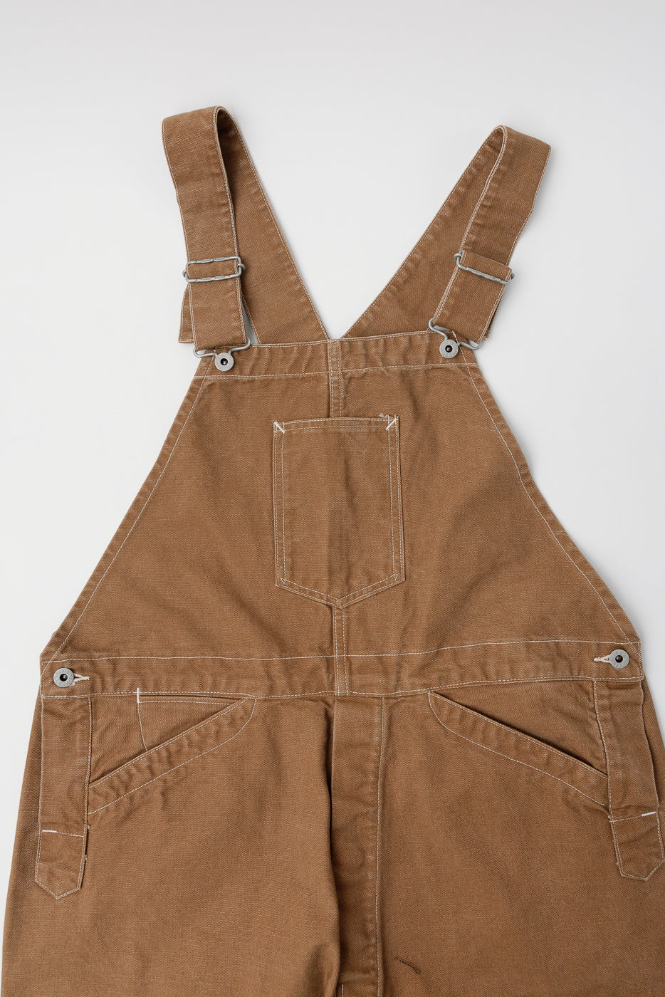 Flat lay close-up of the back upper half of POST O’ALLS brown canvas overalls, showing back yoke, pocket, and strap hardware details.