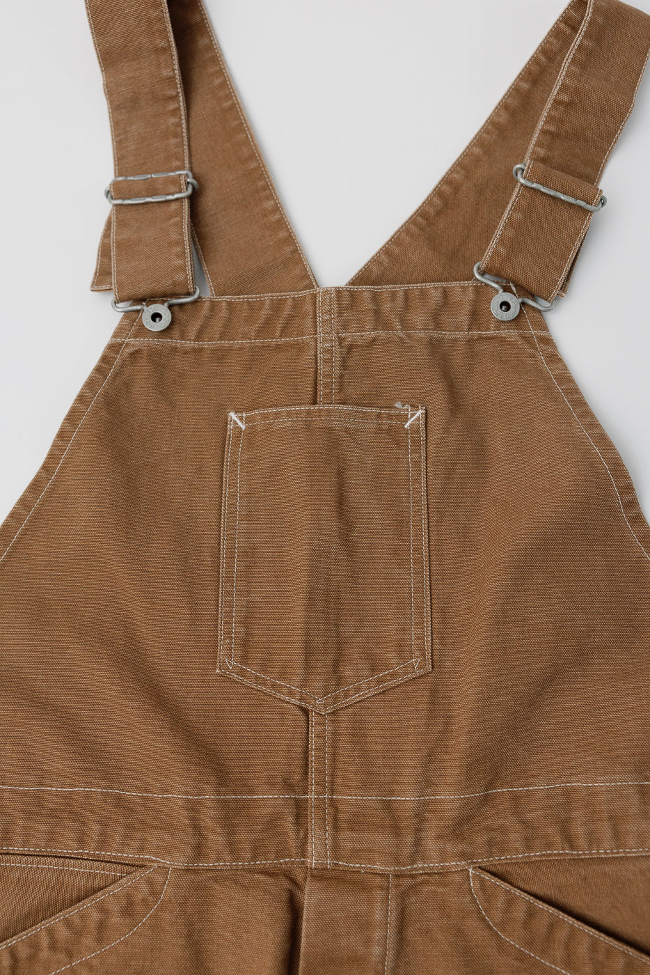 Close-up front view of brown canvas overalls bib, highlighting chest pocket and straps.