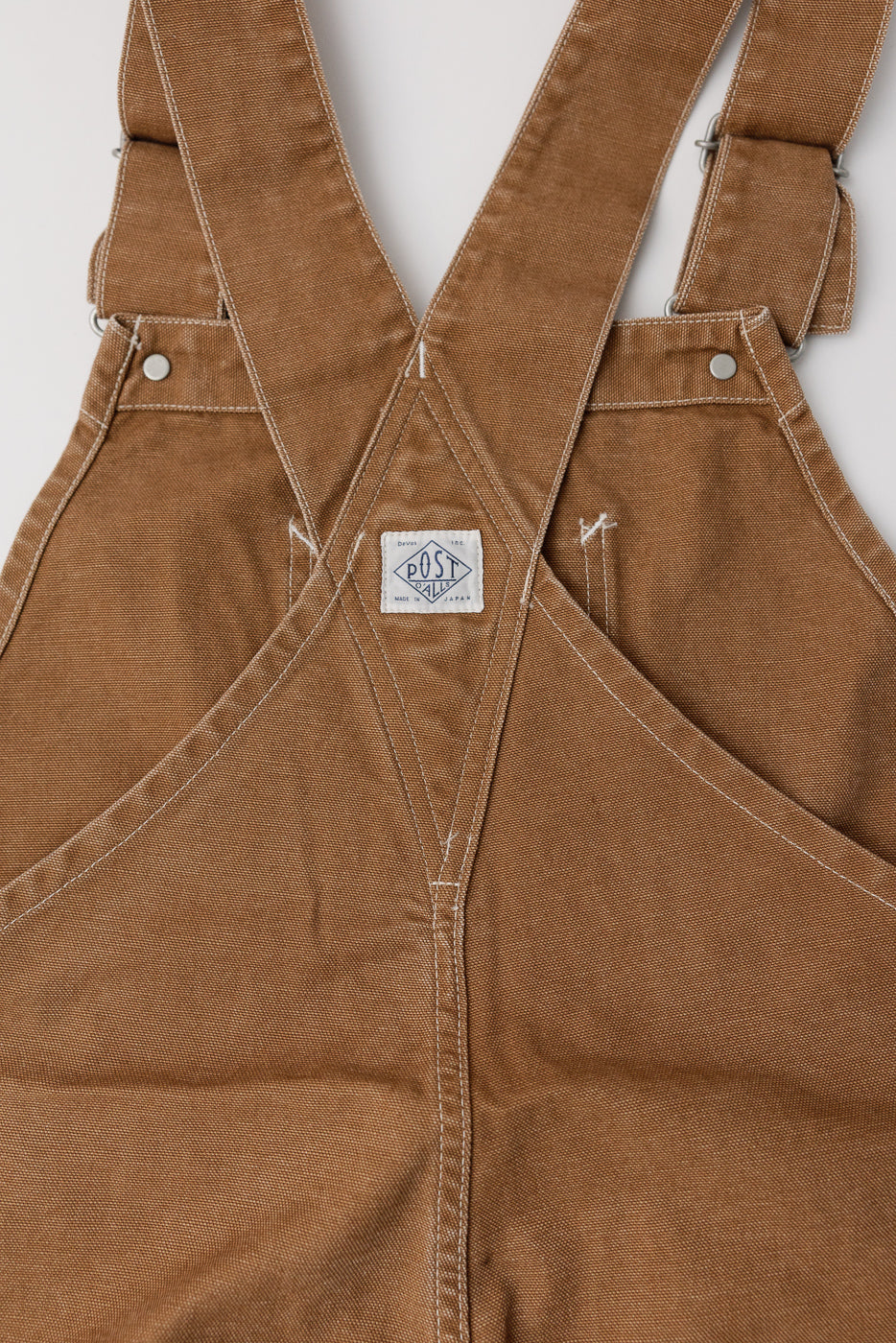 Close-up of back upper section of brown washed canvas overalls, showing POST O’ALLS logo tag, white stitching, and metal button details.
