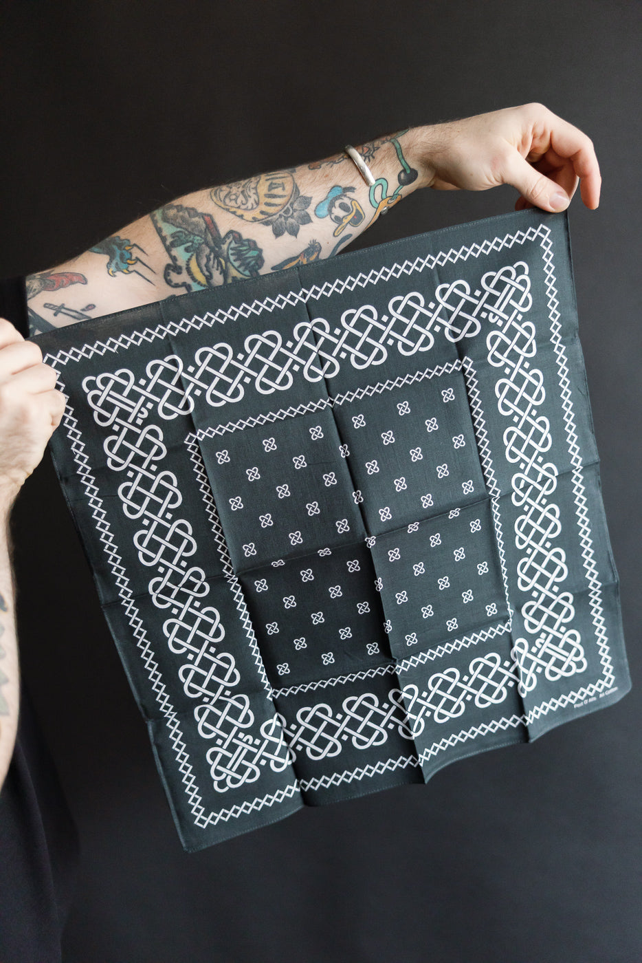 Close-up of Post O'Alls charcoal bandana with rope pattern, front view, held by tattooed male arm