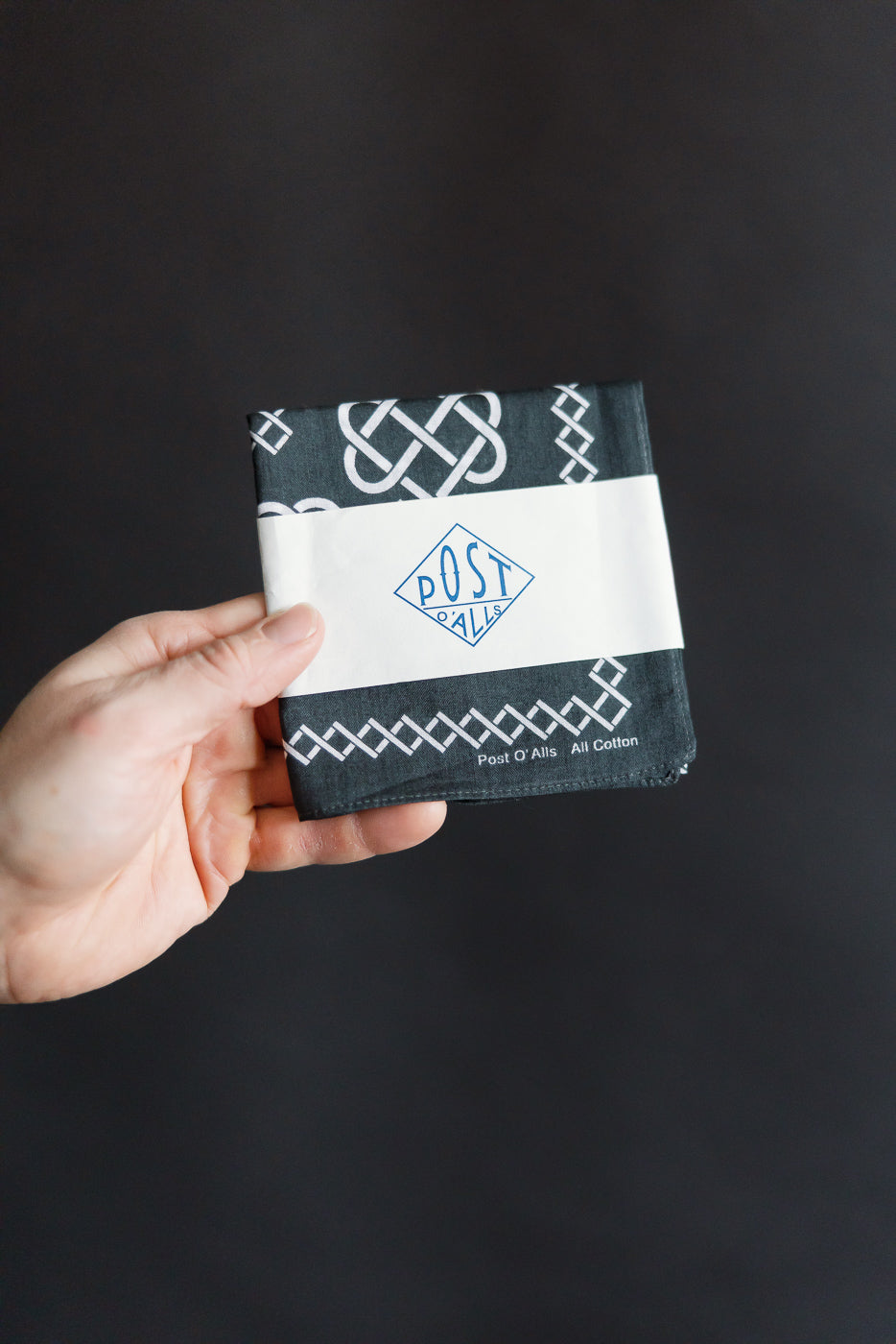 Close-up of hand holding Post O’Alls charcoal bandana, front folded with rope design detail.