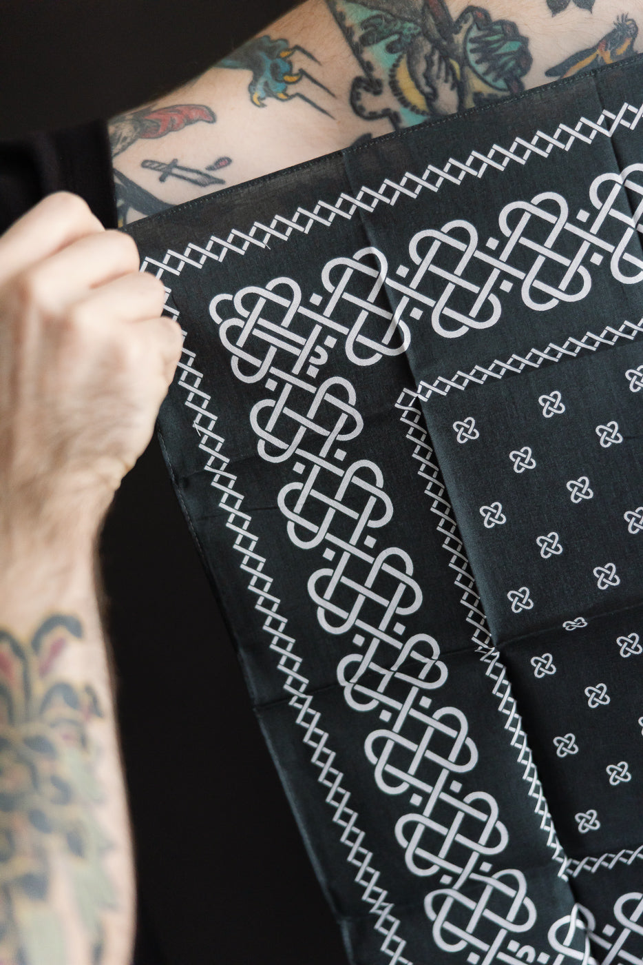 Close-up of Post O’alls charcoal bandana with rope design, detailed border, held by tattooed male model, Dant contemporary accessories.