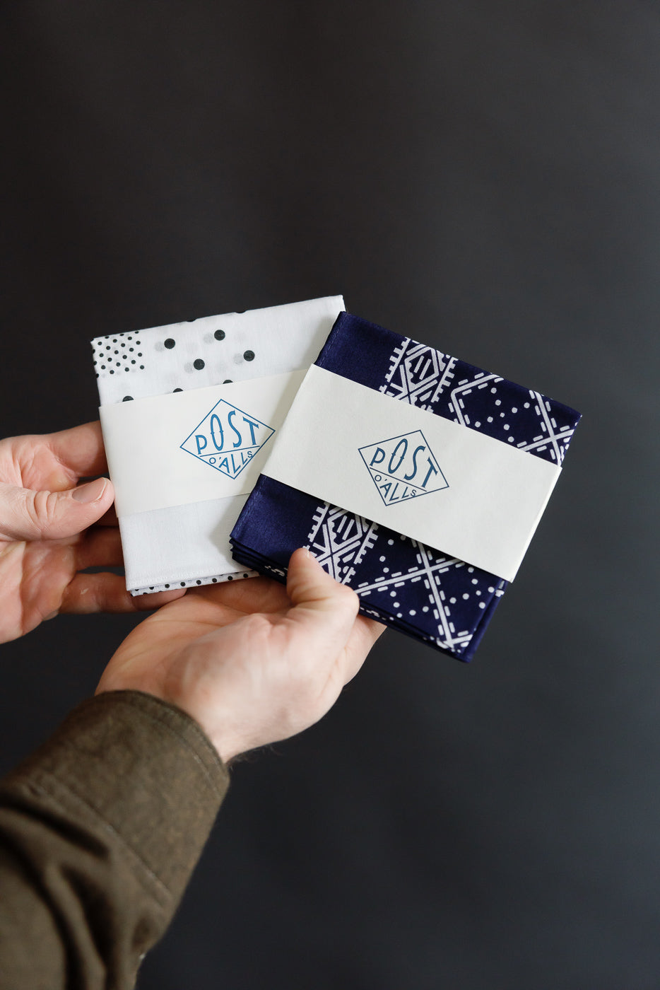 Close-up of hands holding two folded Post O'Alls bandanas, white and navy with stitch design, front view.