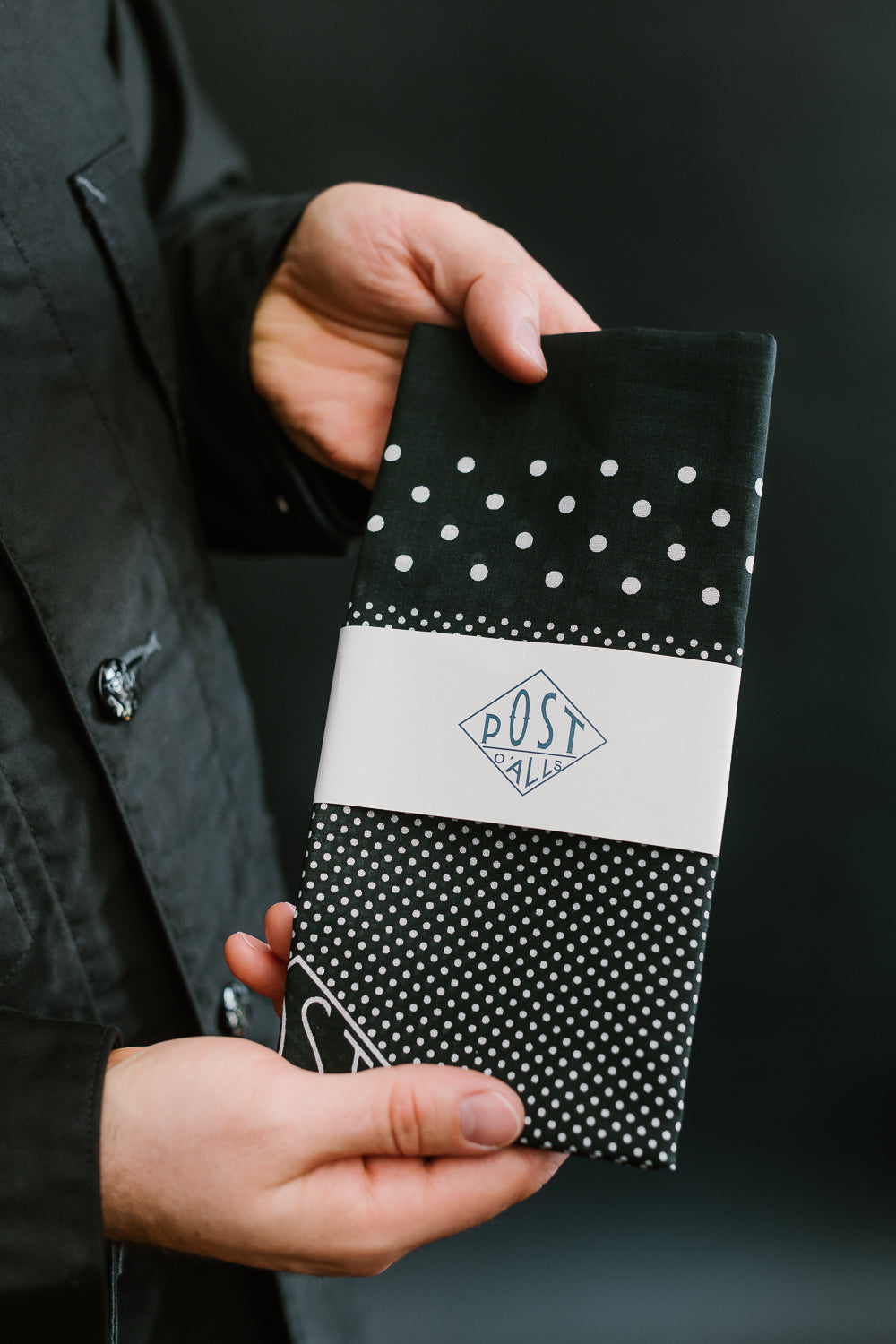 Close-up of male hand holding Post O'Alls charcoal bandana scarf, front polka dot detail