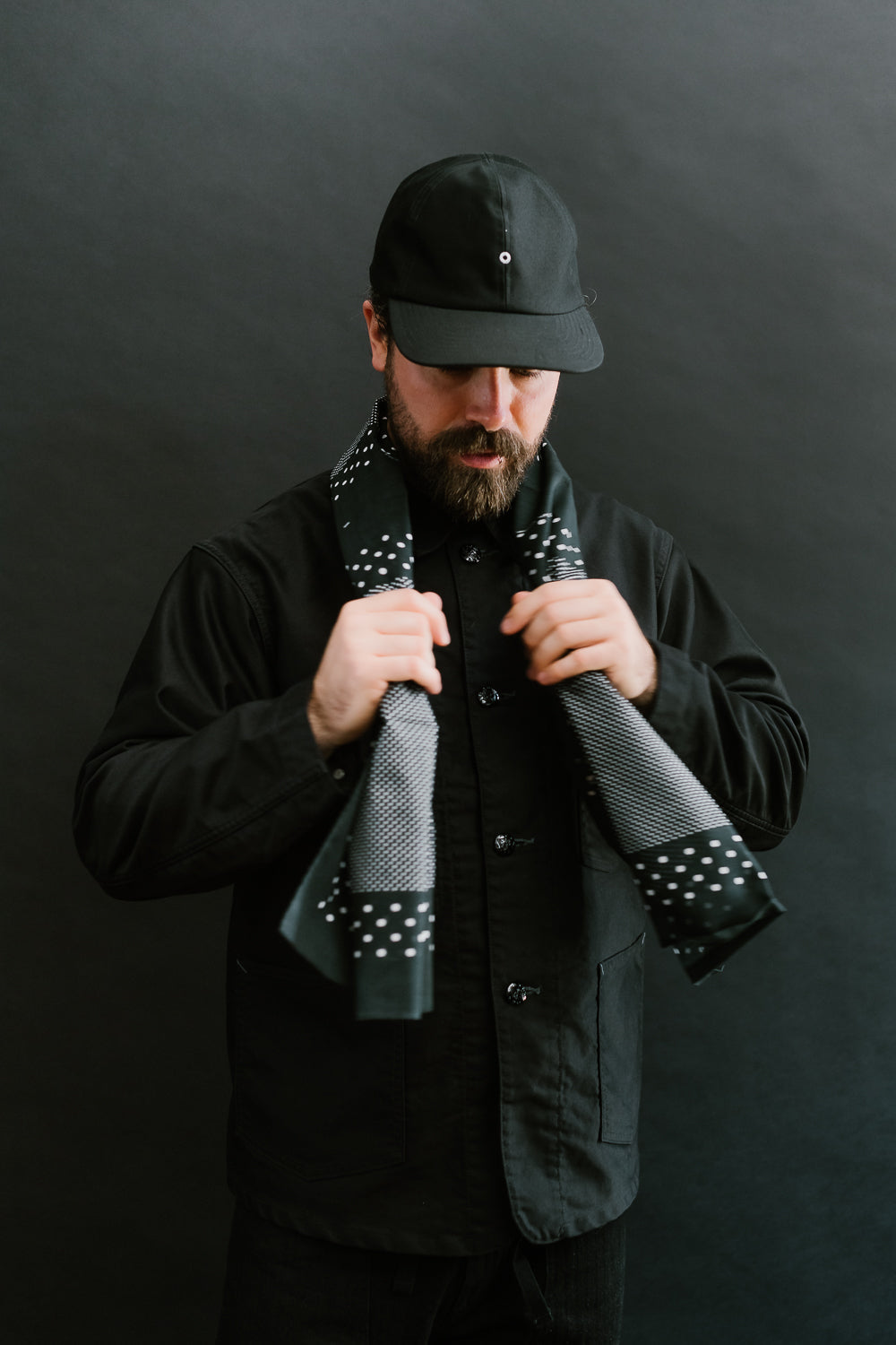 Male model 5'9” 165lbs athletic build in black jacket, front mid-shot, tying charcoal bandana scarf with white dot print, Dant contemporary casual apparel.