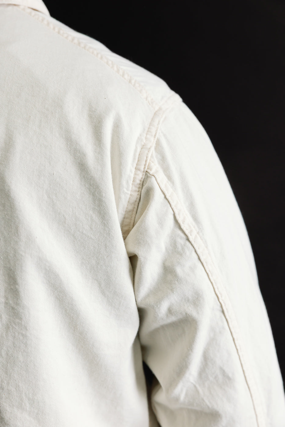 Close-up of the back shoulder seam and sleeve of a natural mid-weight denim jacket, fabric texture visible.
