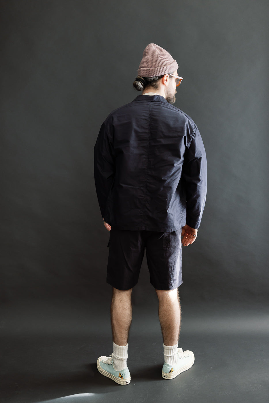 Rear view of a man in the 3135-LMN Post Chinois Travail 2 Light Moleskin jacket, showcasing its cropped, relaxed fit and rounded collar, embodying refined French-Chinese workwear sensibilities, styled with matching shorts.