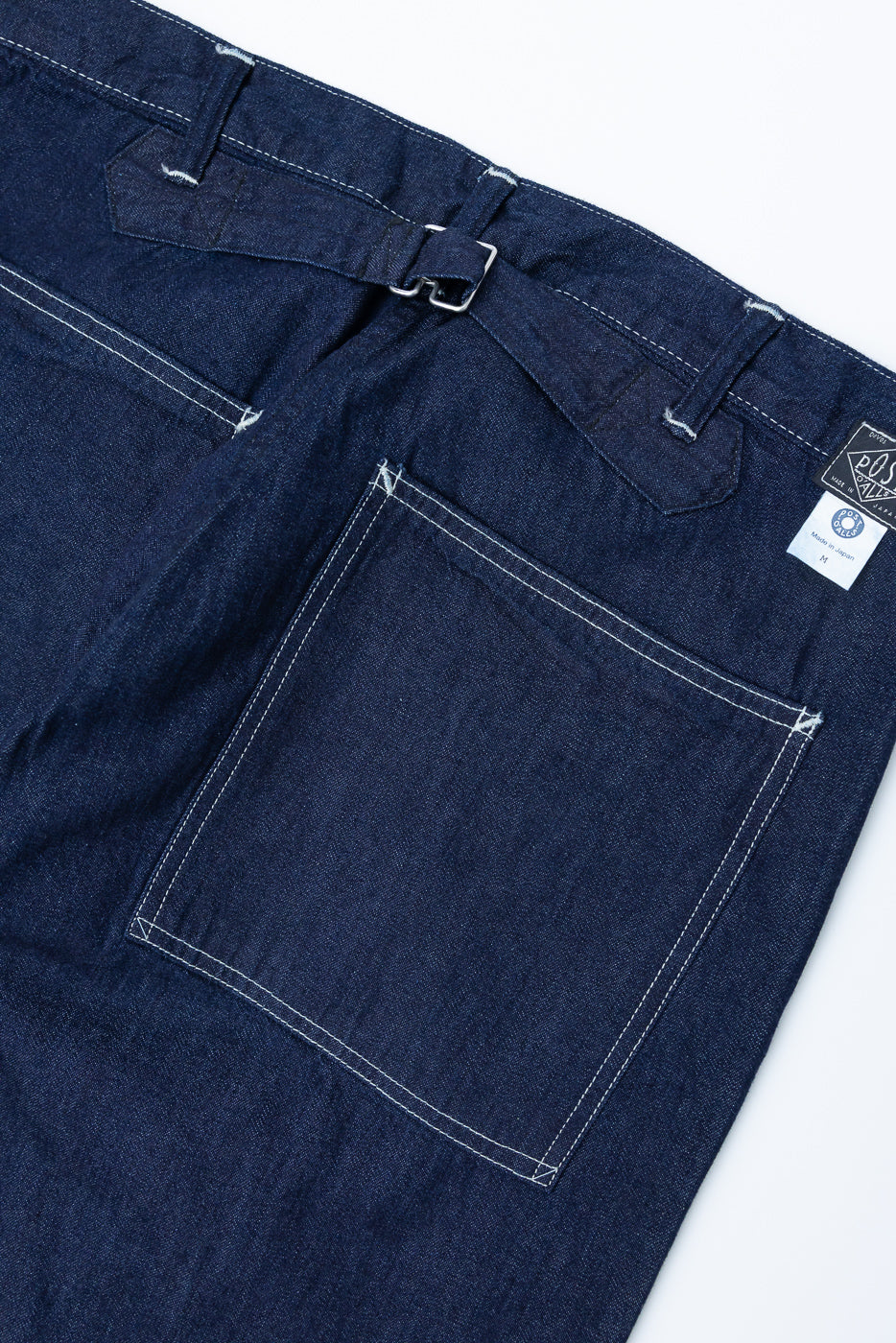 Close-up of the back side of dark indigo denim pants showing rear pockets, buckle adjuster, and contrast stitching.