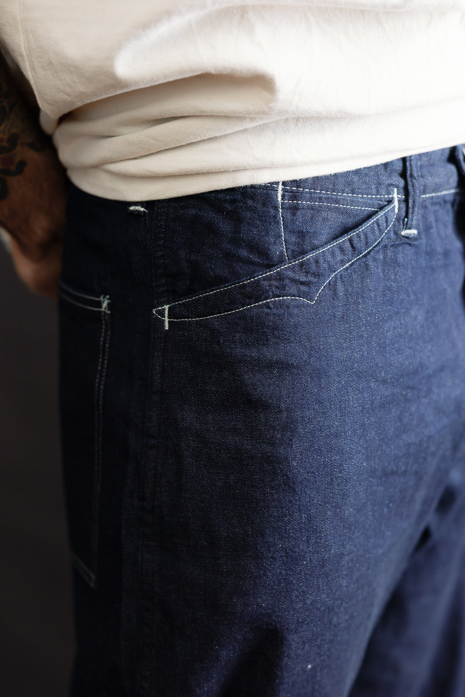 Close-up of men's dark indigo denim jeans left side, showing front pocket and contrast stitching, worn by male model 5'9