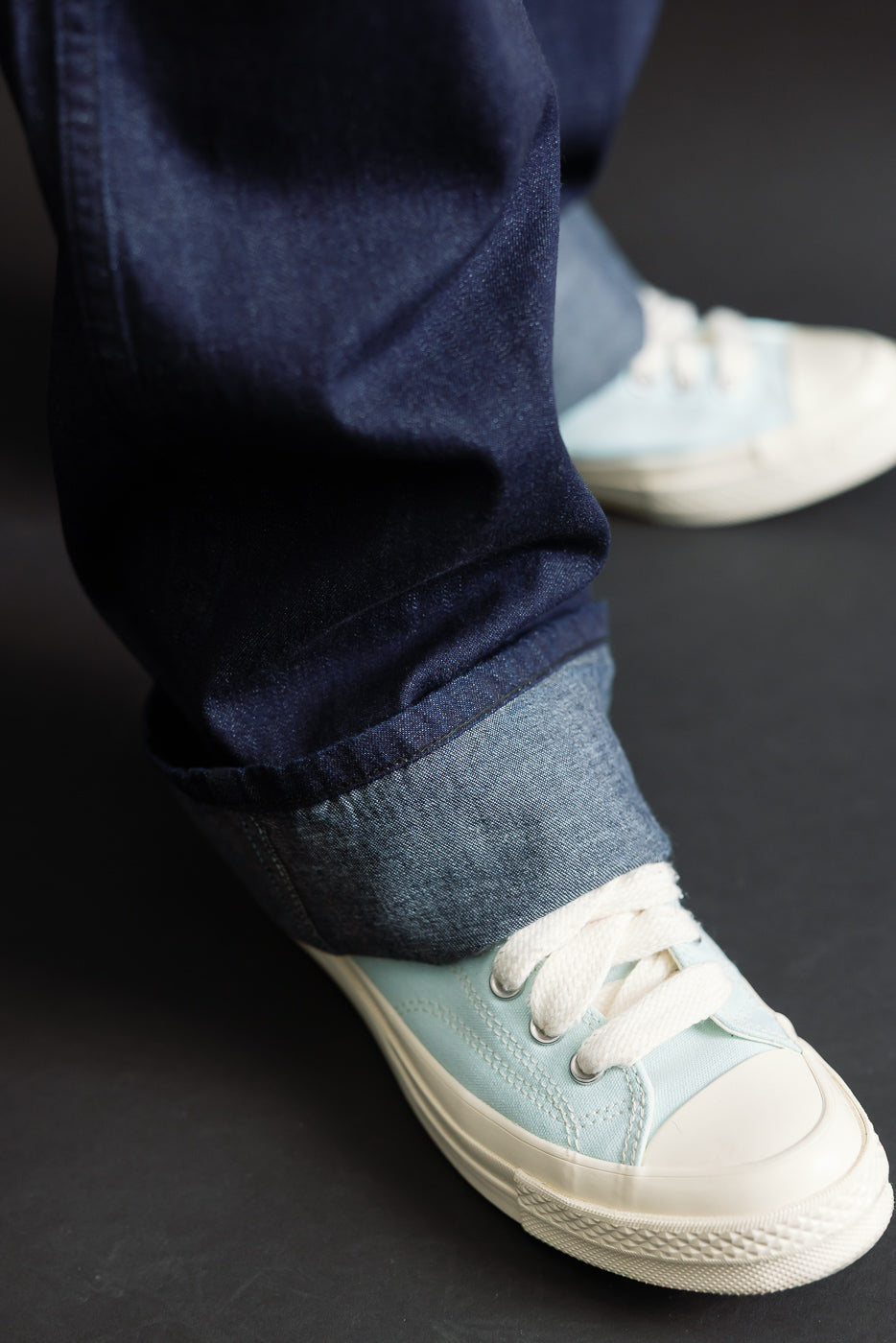 Close-up of male model’s cuffed dark indigo selvedge denim jeans from front, showing hem and fabric texture, paired with light blue casual sneakers. Model is 5'9