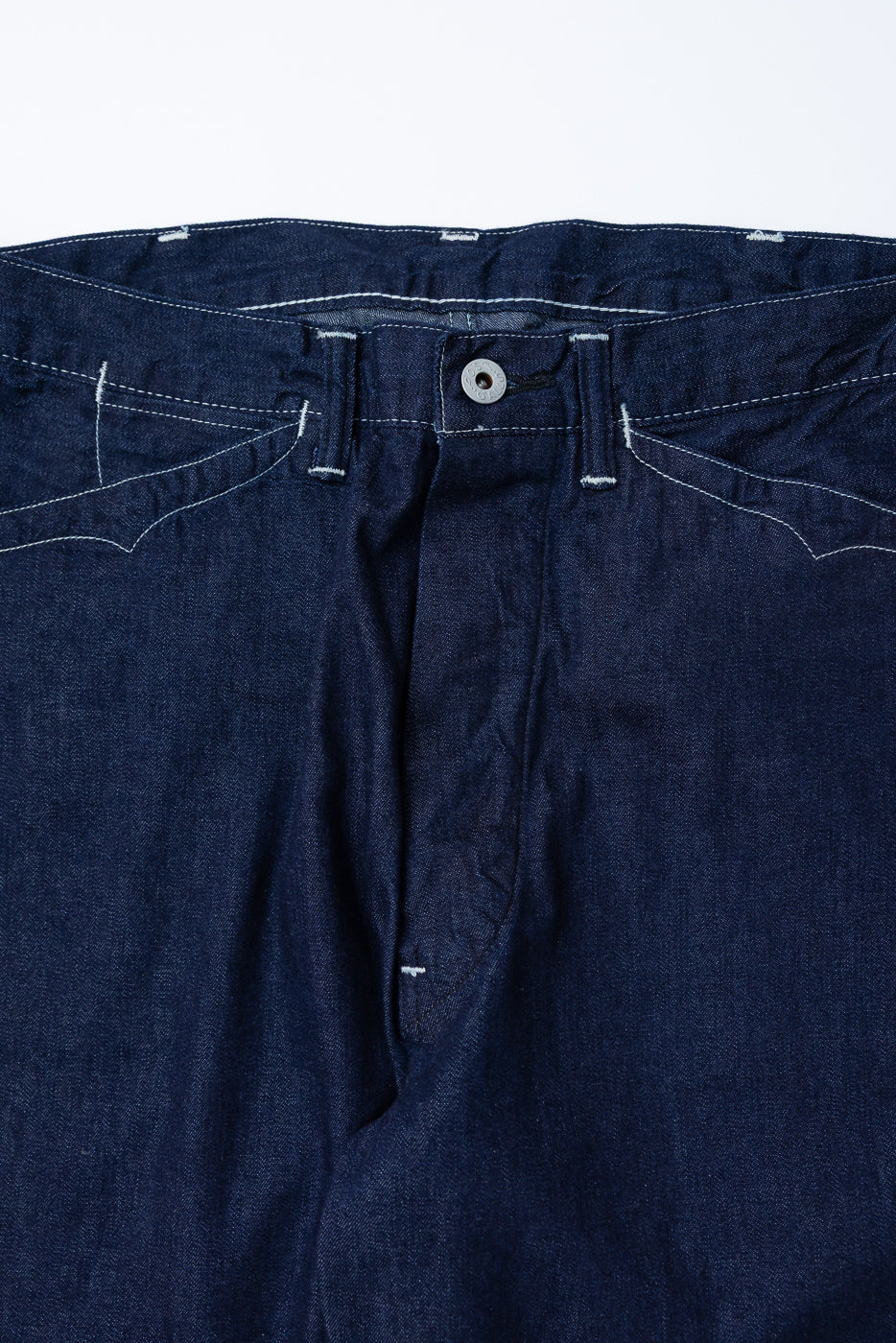Close-up front view of dark indigo denim pants waistband, button, and pocket stitching.