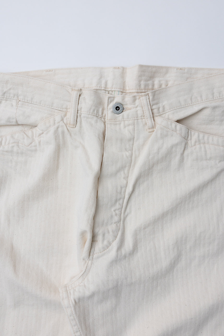Close-up of front waistband and fly of natural herringbone twill pants, showing button and pocket detail.