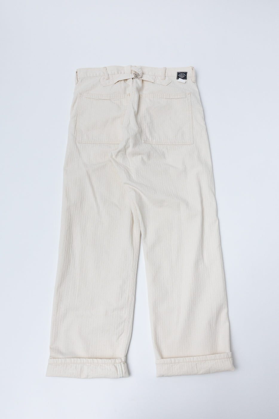 Flat lay of natural herringbone twill pants, full back view, shows pockets and adjustable waist tab.