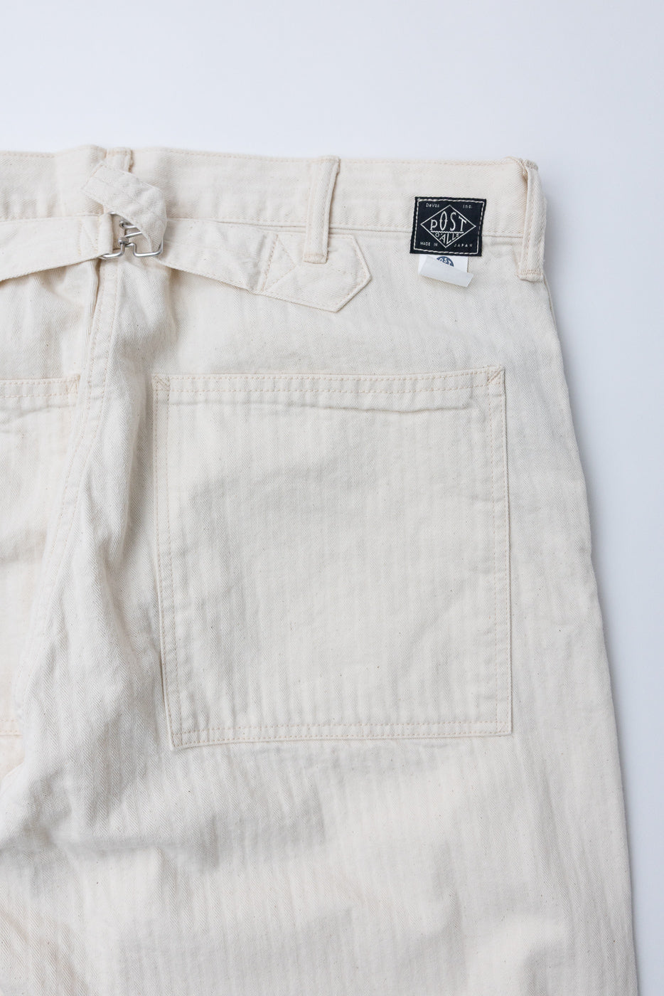 Close-up of back side of natural herringbone twill pants, showing back pocket, belt detail, and fabric texture.