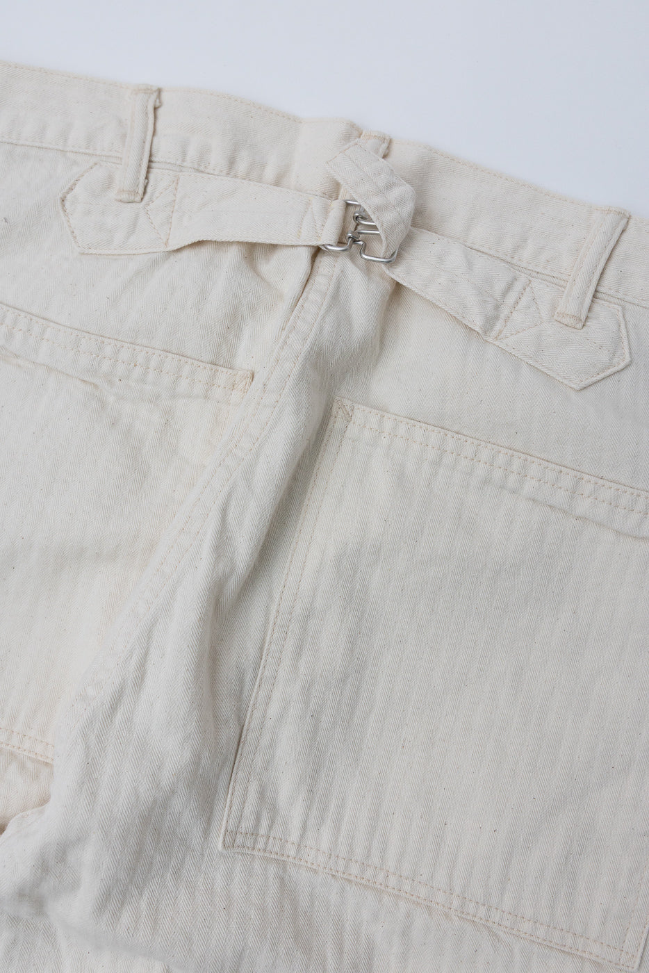 Close-up of the back of natural herringbone twill pants showing rear pockets and buckle cinch detail.