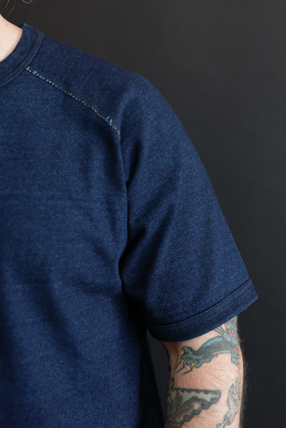 Close-up front view of male model's shoulder & sleeve, showing indigo selvedge tee with contrast stitching, ribbed hem