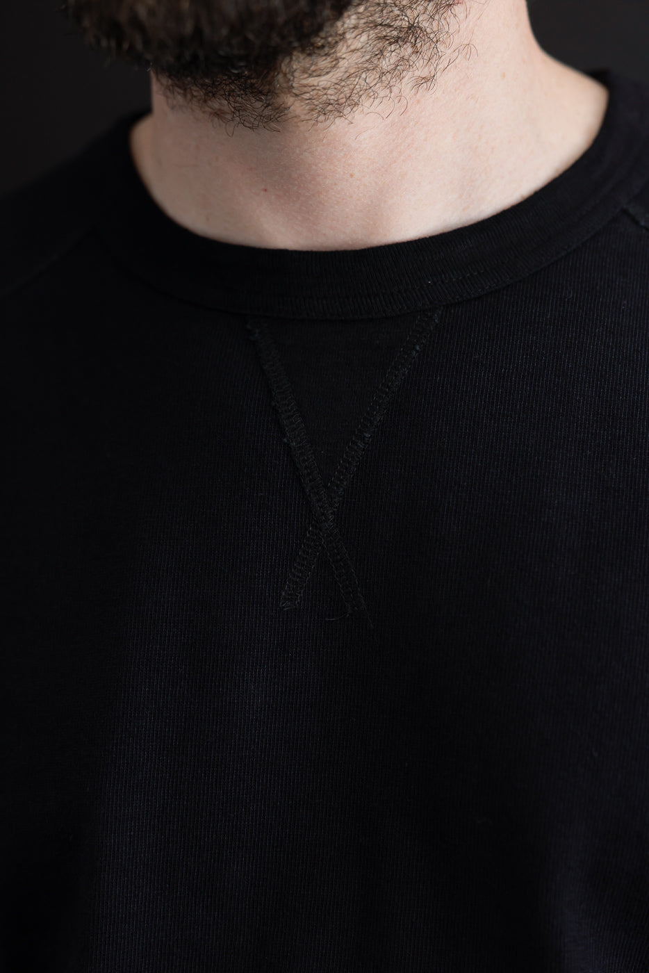 Close-up front view of black crewneck sweatshirt, showcasing ribbed collar and shoulder seam detail on male model.