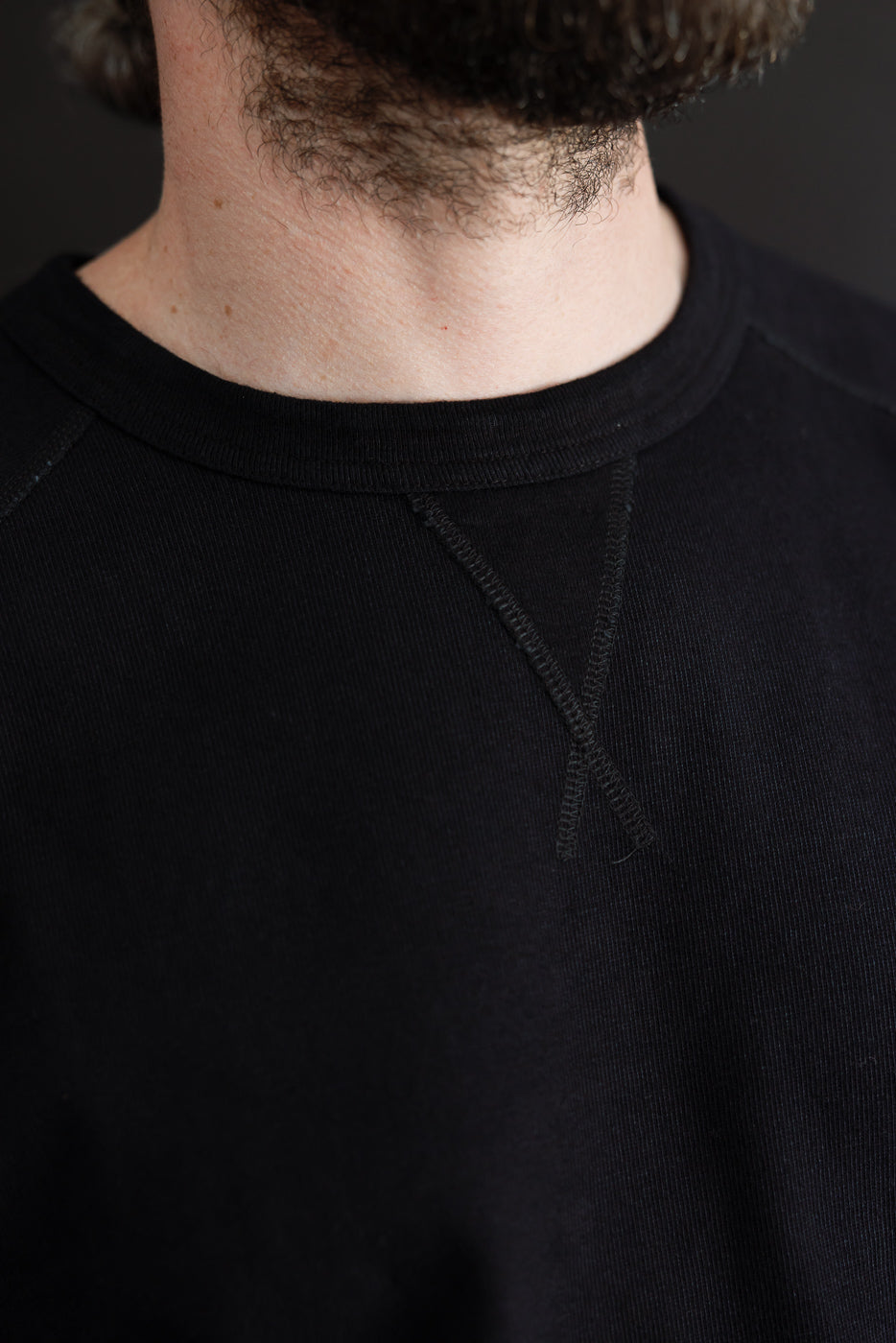 Close-up front view of black crewneck sweatshirt on male model, showing ribbed collar and raglan seam detail