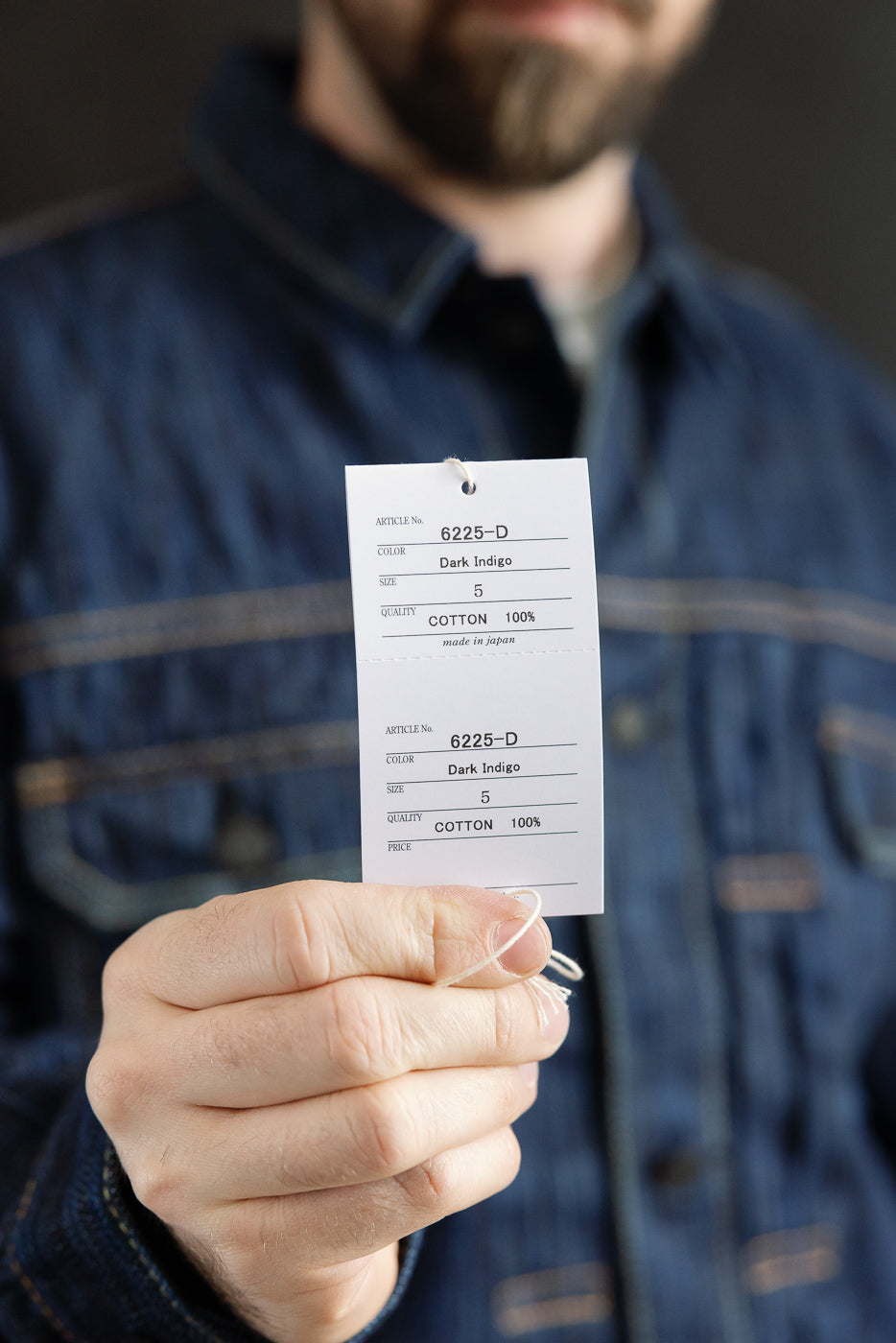 Close-up of a hand holding a white tag on a dark indigo denim jacket, displaying Article No. 6225-D - 14.5oz 'Dark Waterfall' Selvedge Type 2 Jacket - Indigo, highlighting its premium Japanese craftsmanship.