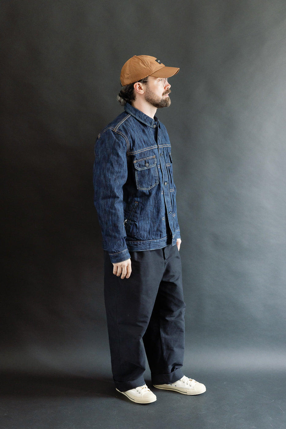 Man in profile wearing the 6225-D - 14.5oz Dark Waterfall Selvedge Type 2 Jacket in Indigo, showcasing its textured denim, button-front, chest flap pockets, and relaxed fit, styled with black pants and off-white sneakers.