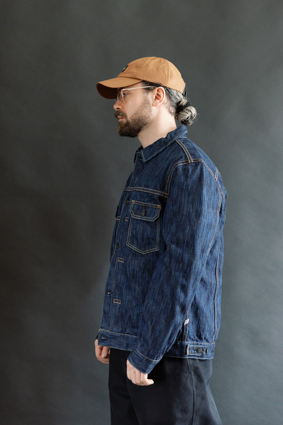 A man models the 6225-D - 14.5oz Dark Waterfall Selvedge Type 2 Jacket, showcasing its textured denim surface, clear-framed glasses, and brown baseball cap, highlighting the jacket's distinctive selvedge detailing and streamlined fit.