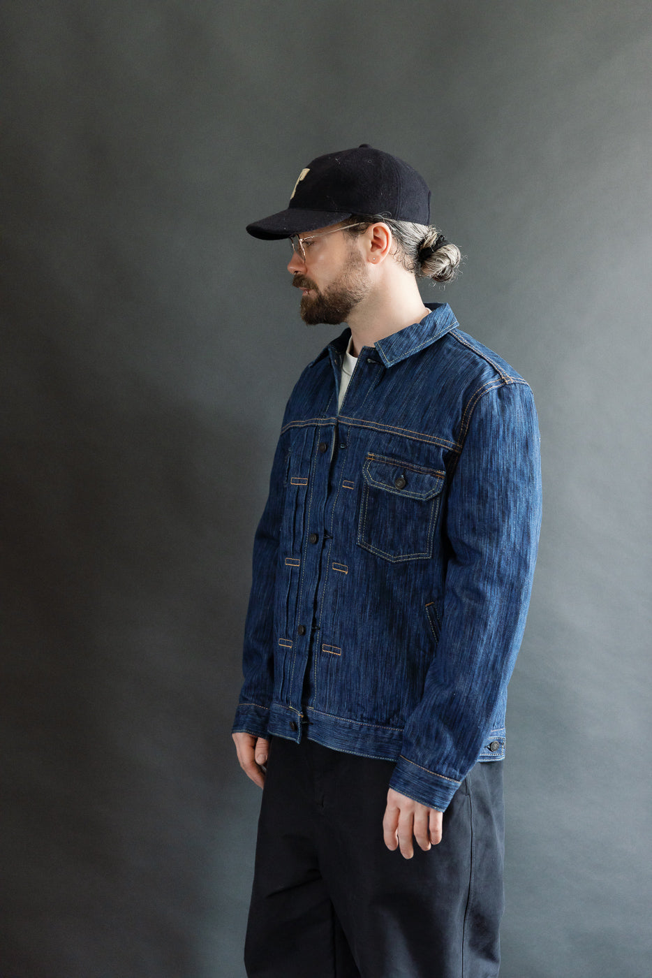 Profile view of a bearded man wearing the 6225-L - 14.5oz Light Waterfall Selvedge Type II Jacket - Indigo, showcasing its denim texture, chest pocket, and modern fit against a gray background.