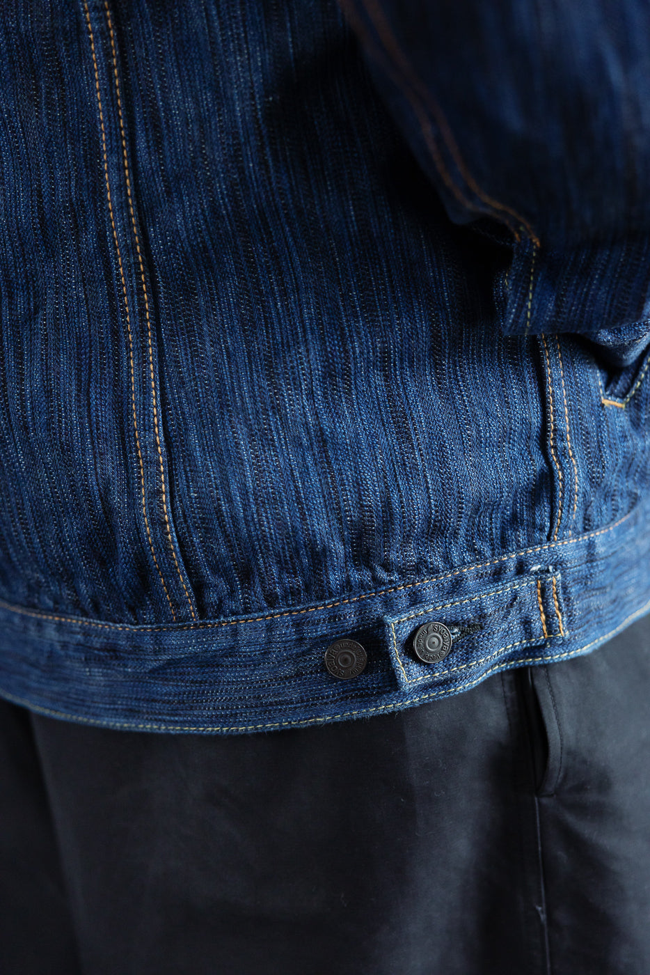 Close-up of the 6225-L - 14.5oz Light Waterfall Selvedge Type II Jacket's lower back, showcasing blue denim's vertical weave, orange stitching, and two metal adjustment buttons over dark pants.