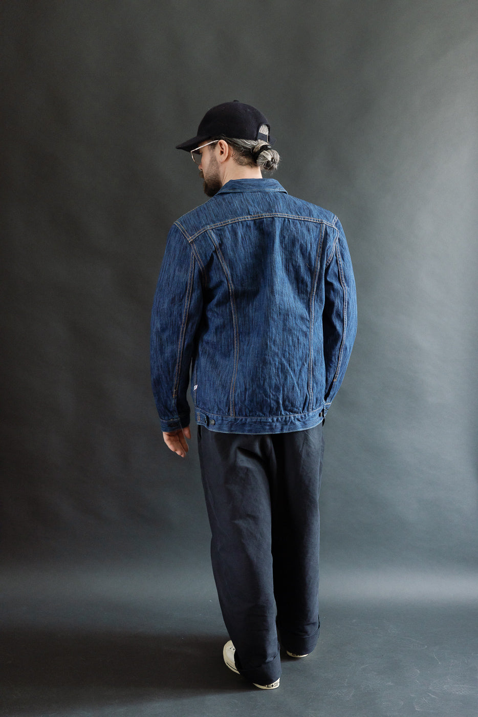 A man models the 6225-L - 14.5oz Light Waterfall Selvedge Type II Jacket in indigo, showcasing its textured Japanese denim, streamlined fit, and signature detailing against a simple gray backdrop.