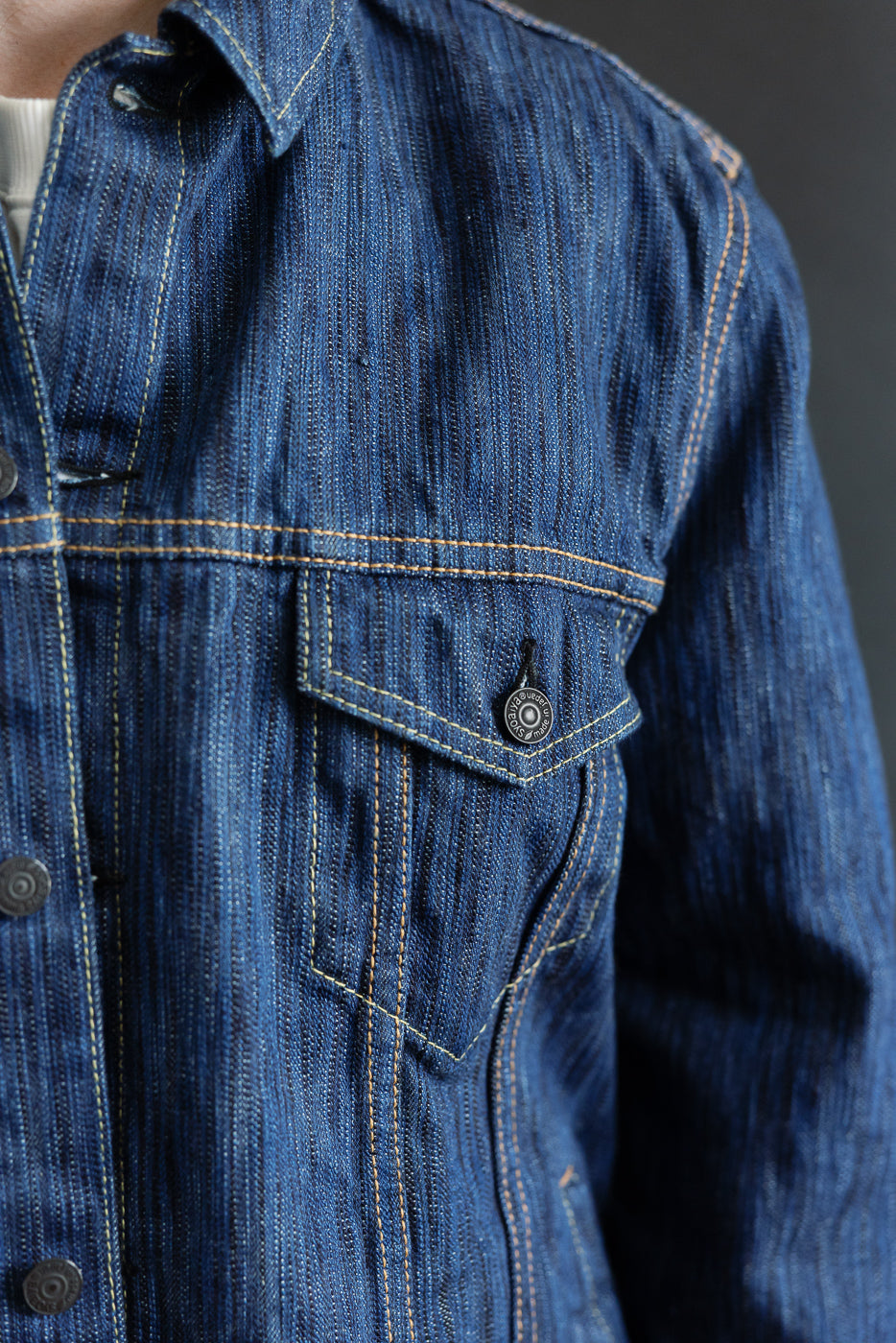 Close-up of the 6226-L - 14.5oz Light Waterfall Selvedge Type III Jacket in Indigo, showcasing vertical slub texture, golden topstitching, flap pocket with metal snap, collar, and front-button placket over a white T-shirt.