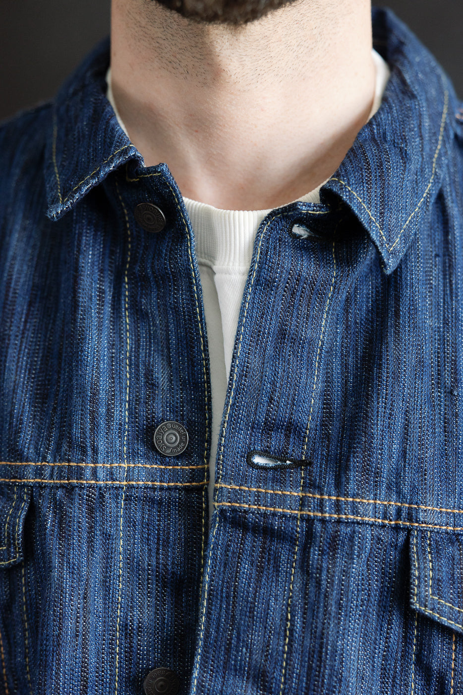 Close-up of the 6226-L - 14.5oz Light Waterfall Selvedge Type III Jacket - Indigo, highlighting its textured Japanese selvedge denim, contrast yellow stitching, and metal snap buttons against a white rib-knit crewneck.
