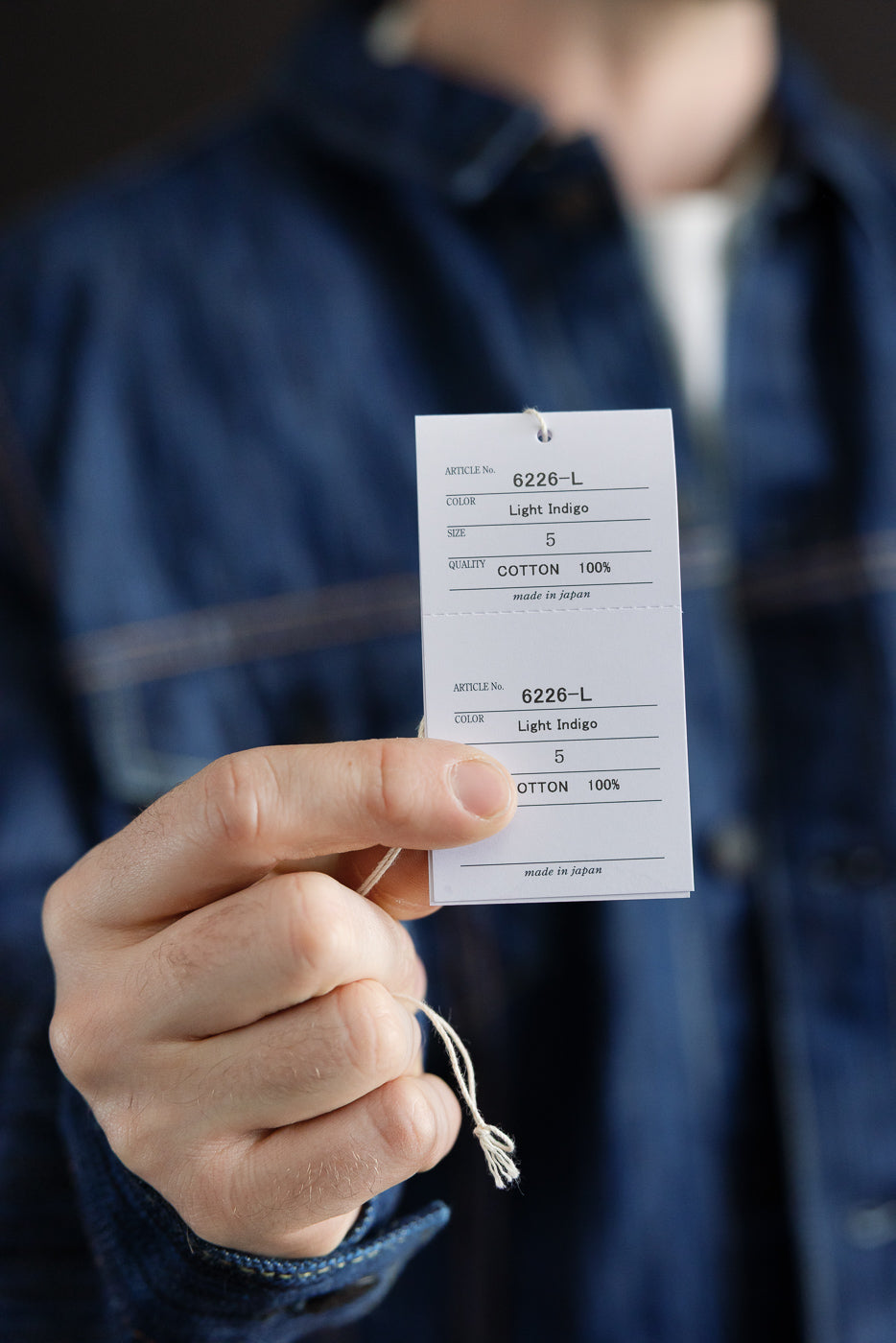 Close-up of hands holding two white tags for the 6226-L - 14.5oz Light Waterfall Selvedge Type III Jacket - Indigo, showcasing product details and emphasizing its Japanese craftsmanship and textured denim quality.