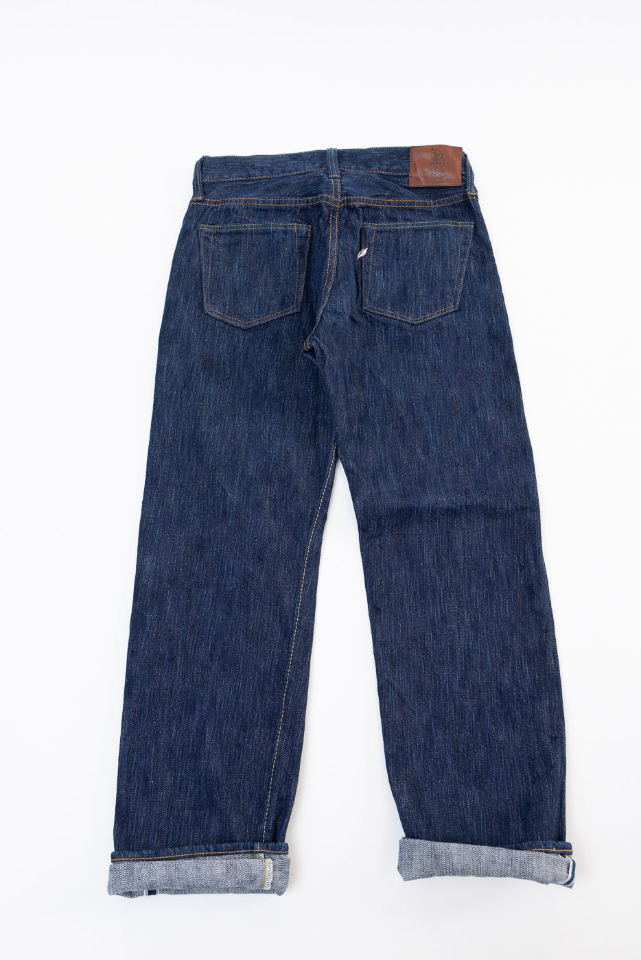 Back view flat lay of dark indigo selvedge denim jeans with leather patch and cuffed hems.