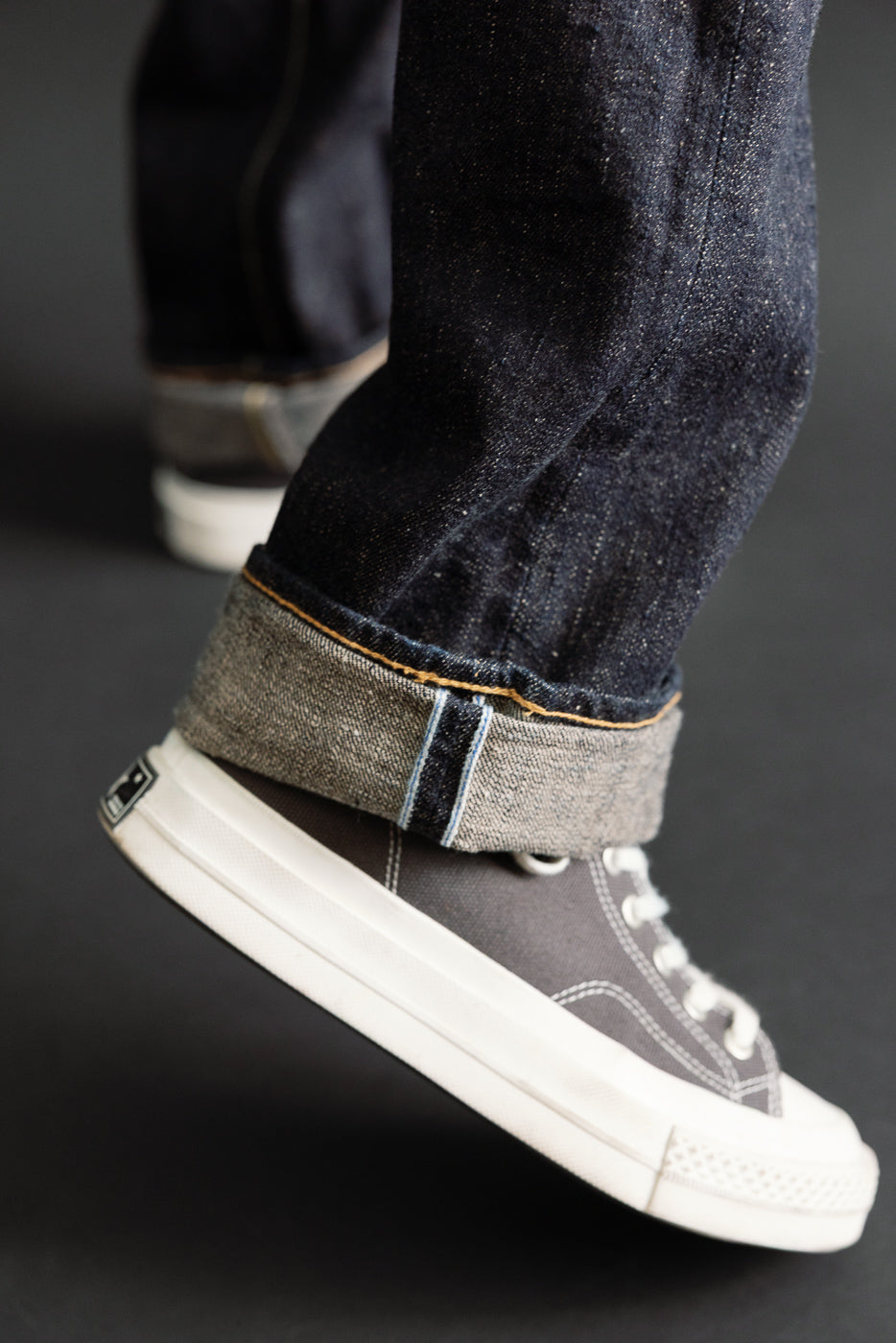 Close-up of the cuffed hem of dark indigo selvedge denim jeans paired with grey canvas sneakers.