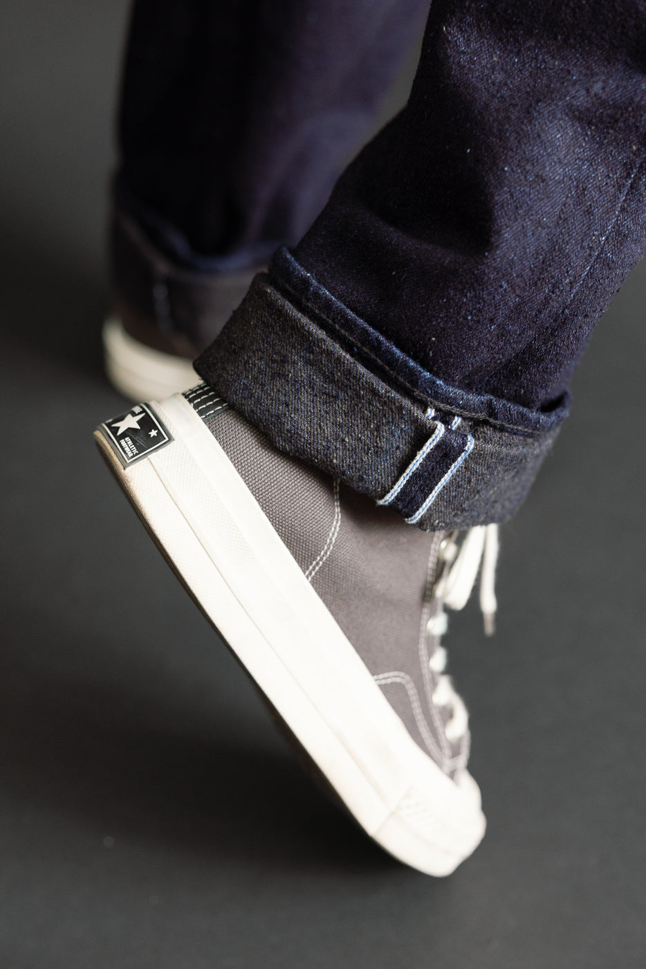 Close-up side view of a male model's feet wearing dark selvedge denim cuffed over grey canvas high-top sneakers.