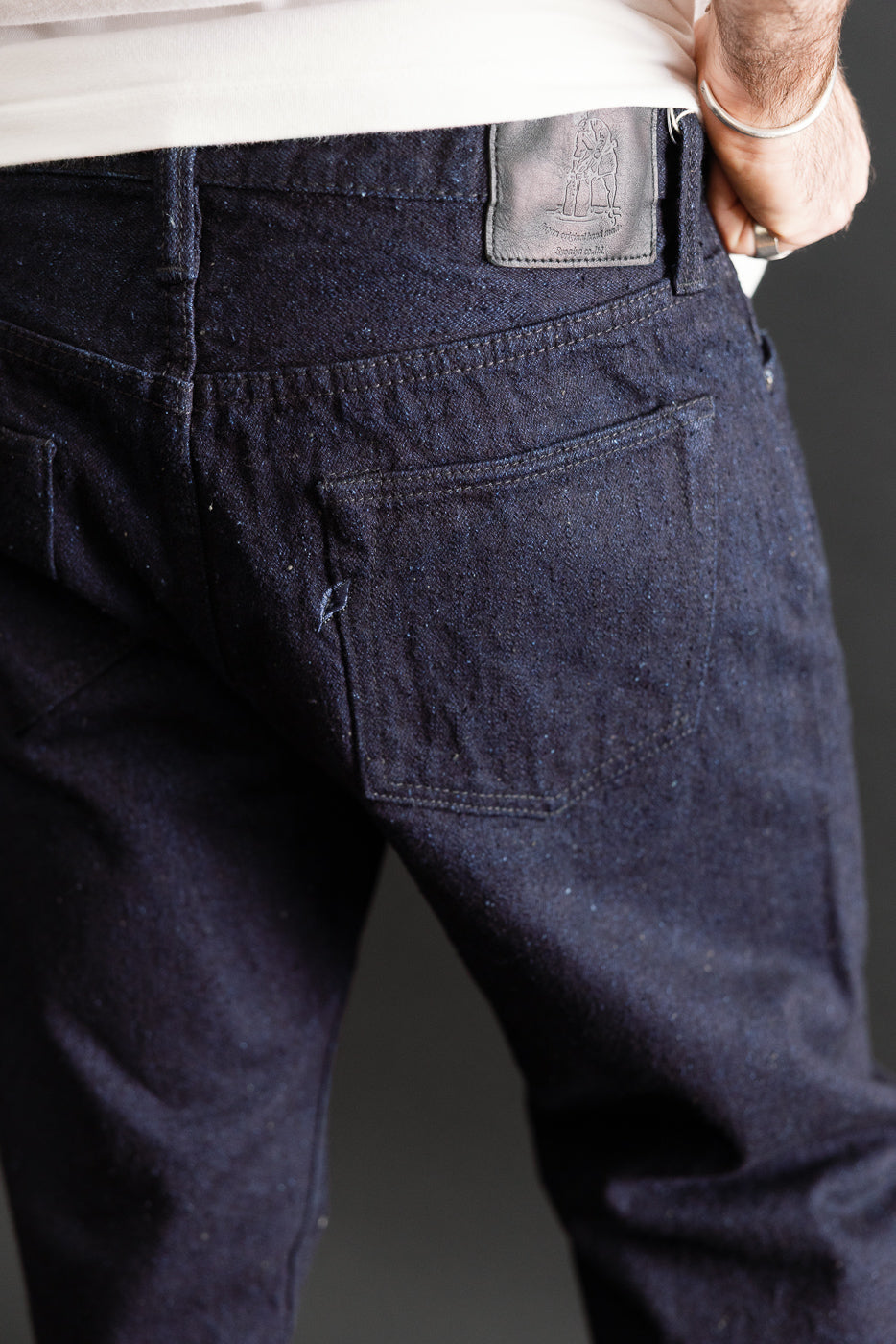 Close-up back view of dark indigo denim jeans showing rear pocket and leather waist patch.