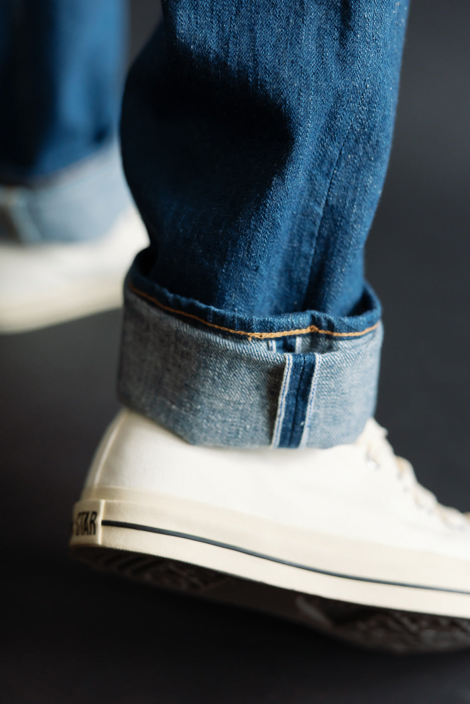 Close-up of selvedge cuff and fabric texture on front leg of Pure Blue Japan greencast indigo jeans, male model, athletic build, with white sneaker.