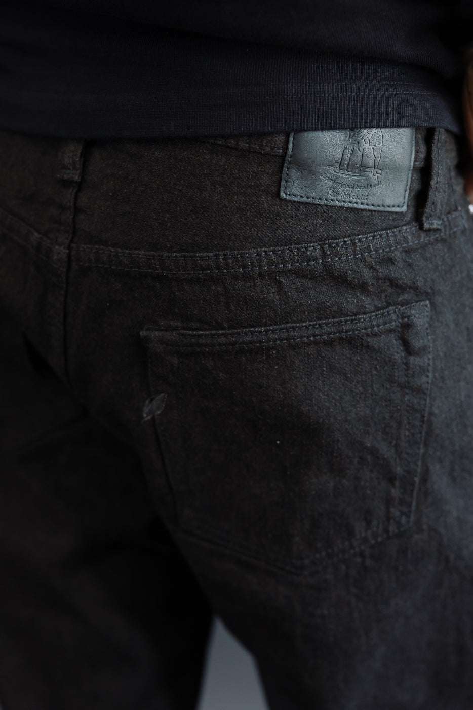 Close-up back view of dark selvedge jeans on male model, showing leather patch, rear pocket, and stitching detail
