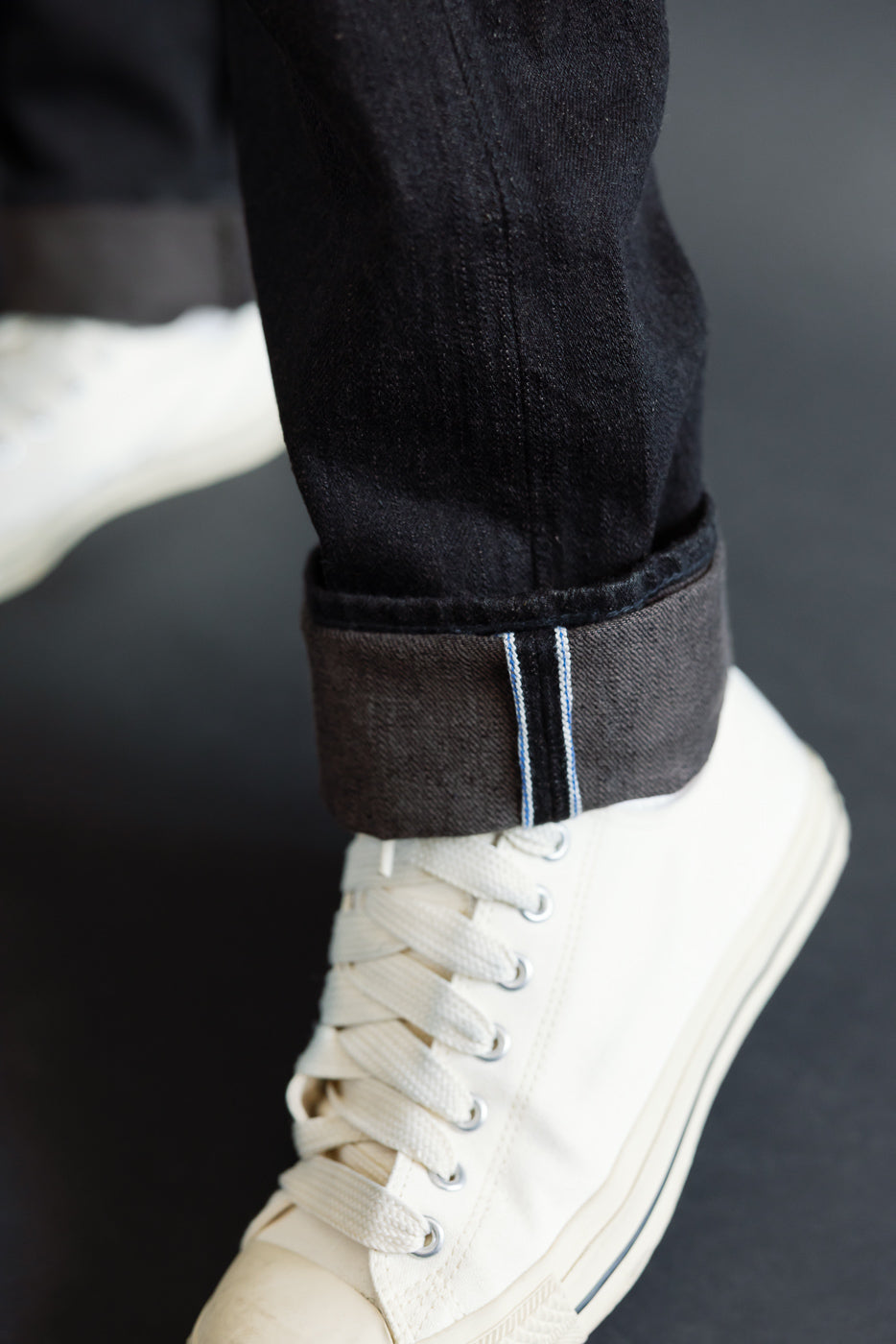 Close-up of male model's cuffed selvedge denim jeans showing raw hem and white sneaker, front view