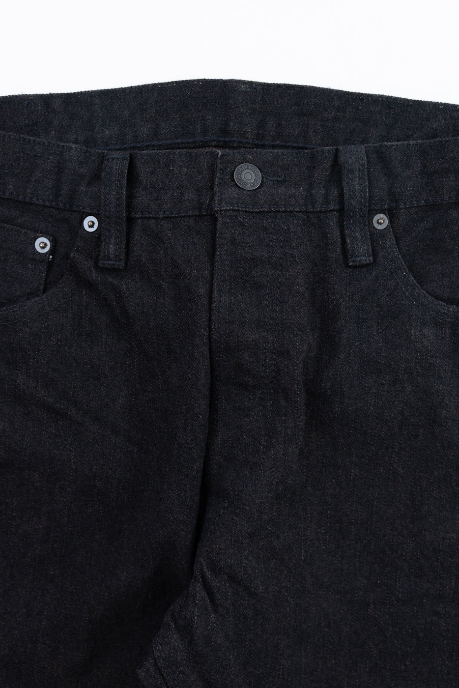Close-up front view of dark selvedge denim jeans, showing button fly, metal rivets, and textured fabric weave.