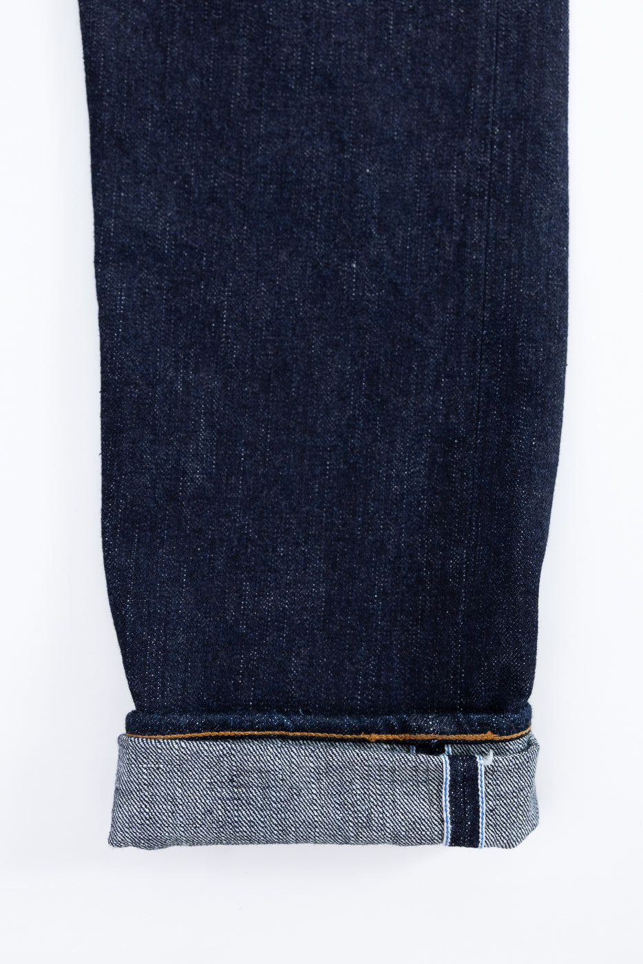 Close-up front view of dark selvedge denim jeans leg, showing raw hem with orange stitching and selvedge edge detail