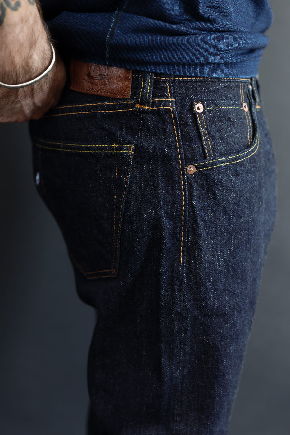 Close-up of male model's back, showcasing dark selvedge jeans with yellow stitching and leather patch.