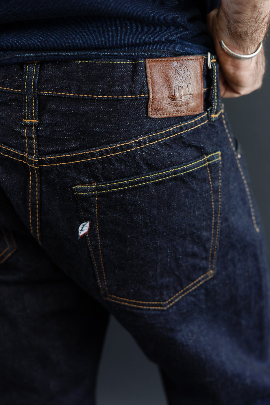 Close-up back view of male model's dark selvedge denim jeans, showing leather patch, yellow stitching, and rear pocket detail