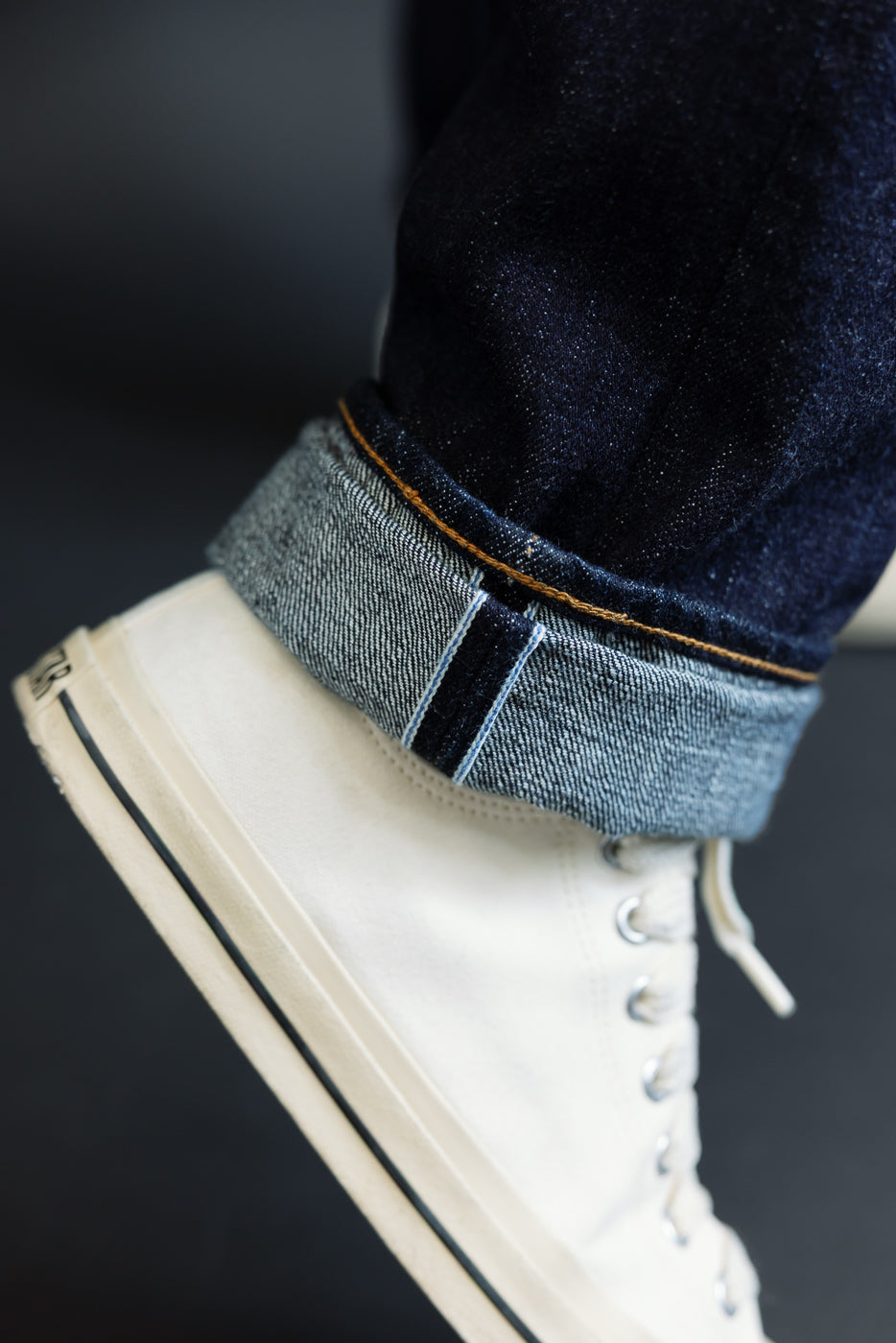 Close-up of male model's cuffed selvedge jeans with orange stitching, paired with white high-top sneakers