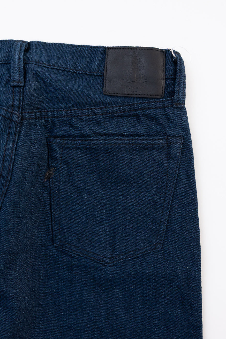 Close-up of the back right side of dark indigo denim jeans showing the back pocket and leather patch.