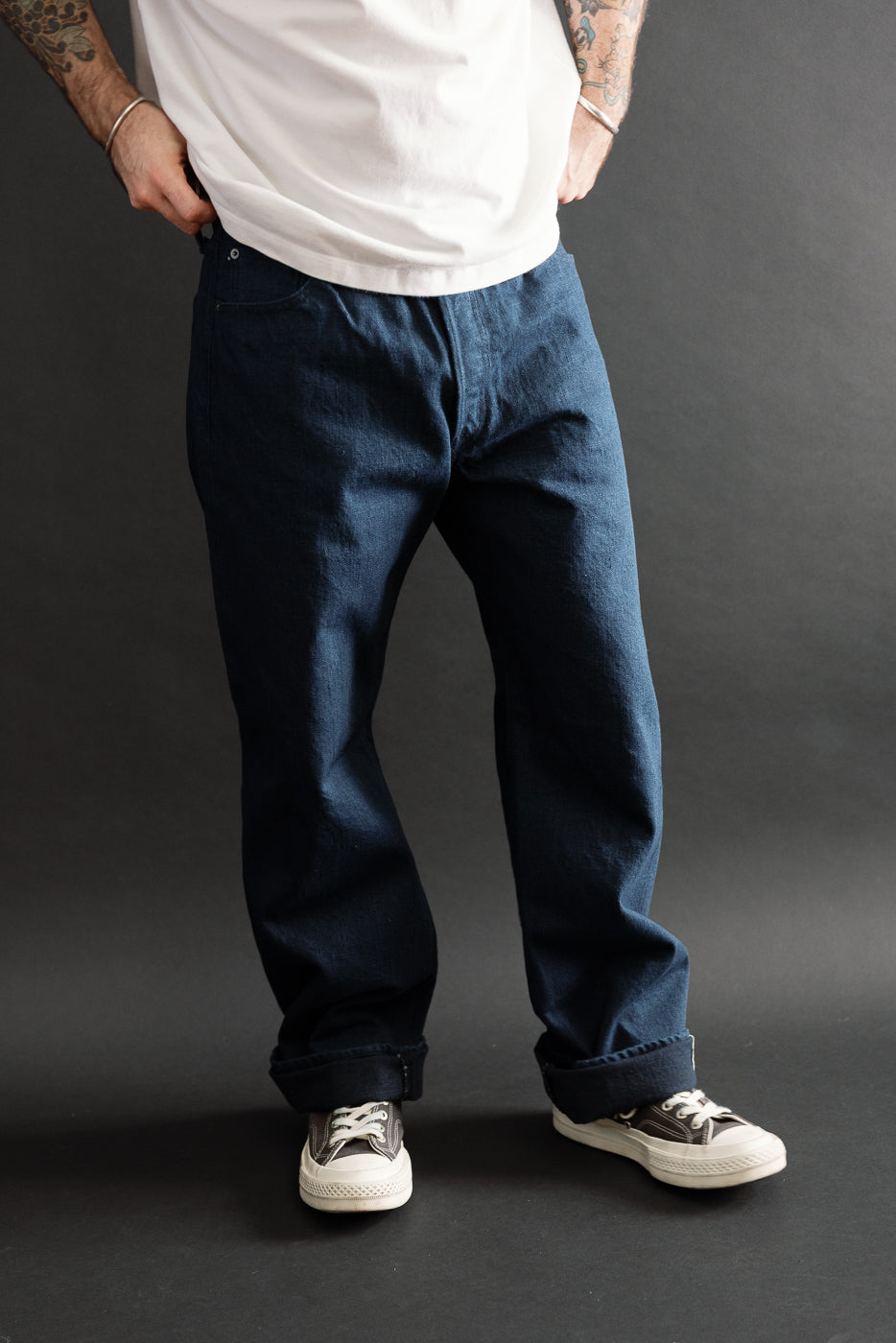 Full body front view of a male model with an athletic build wearing dark blue selvedge denim jeans and black sneakers.