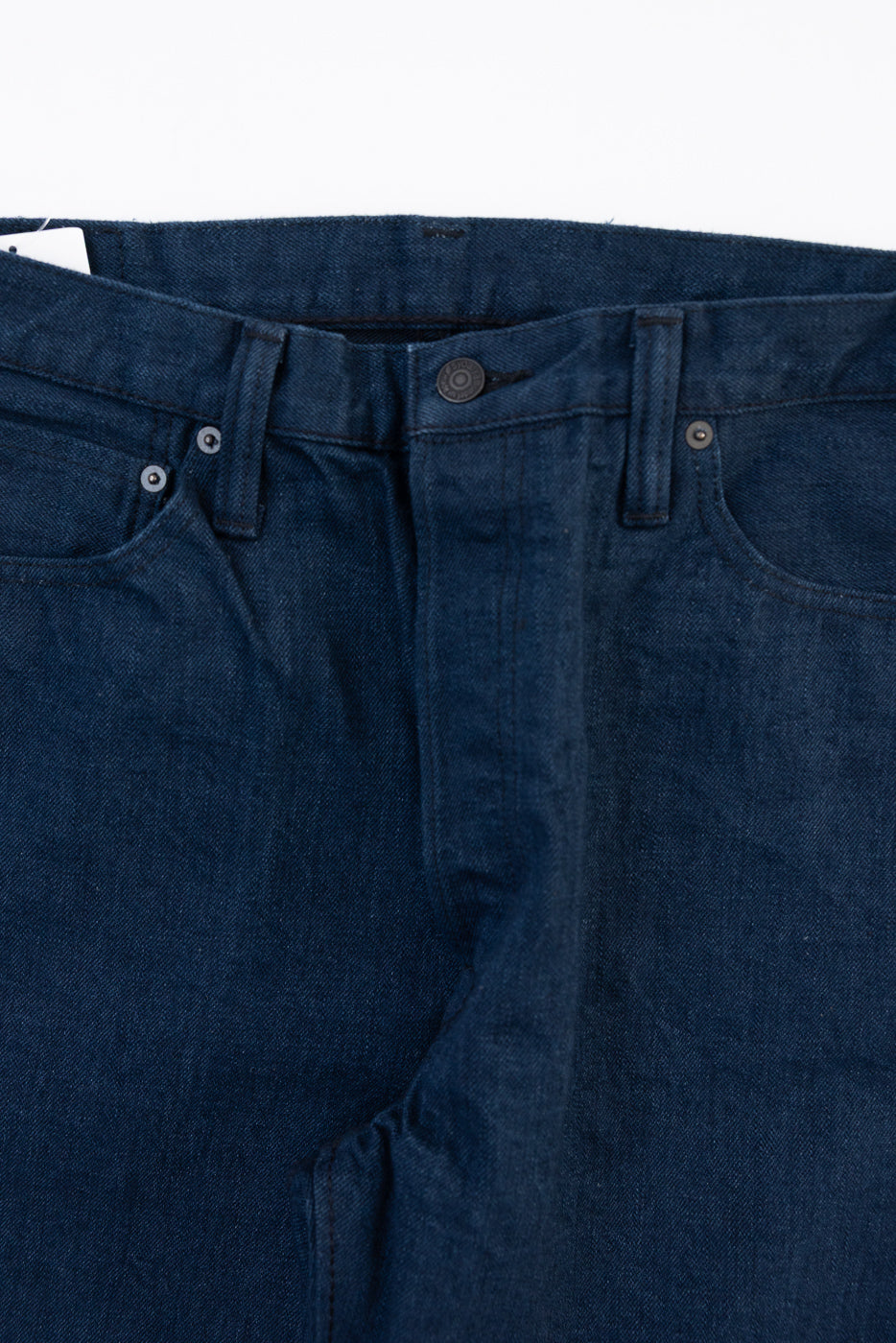 Close-up front view of dark indigo denim jeans showing the waistband, button, and fly texture.