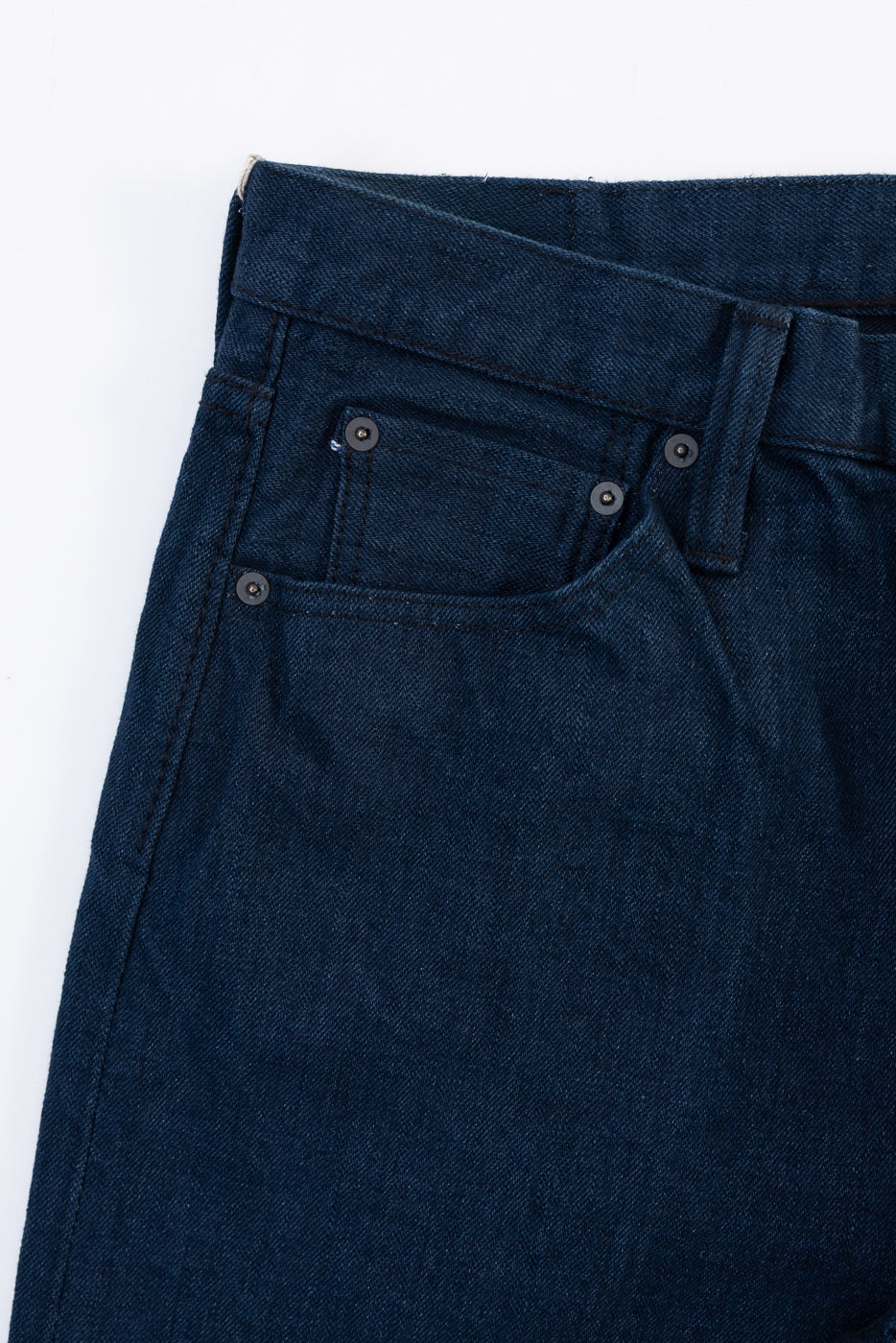 Close-up of the front waist and pocket of dark indigo denim jeans showing fabric texture and rivets.