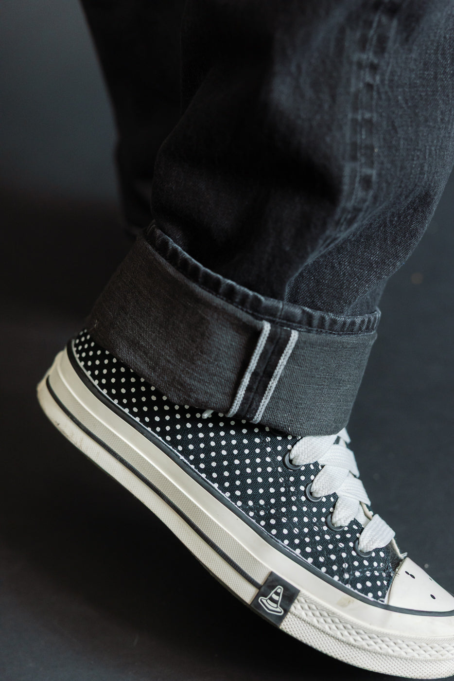 Close-up of male model's cuffed dark denim jeans with white selvedge stripe, paired with polka dot canvas sneakers