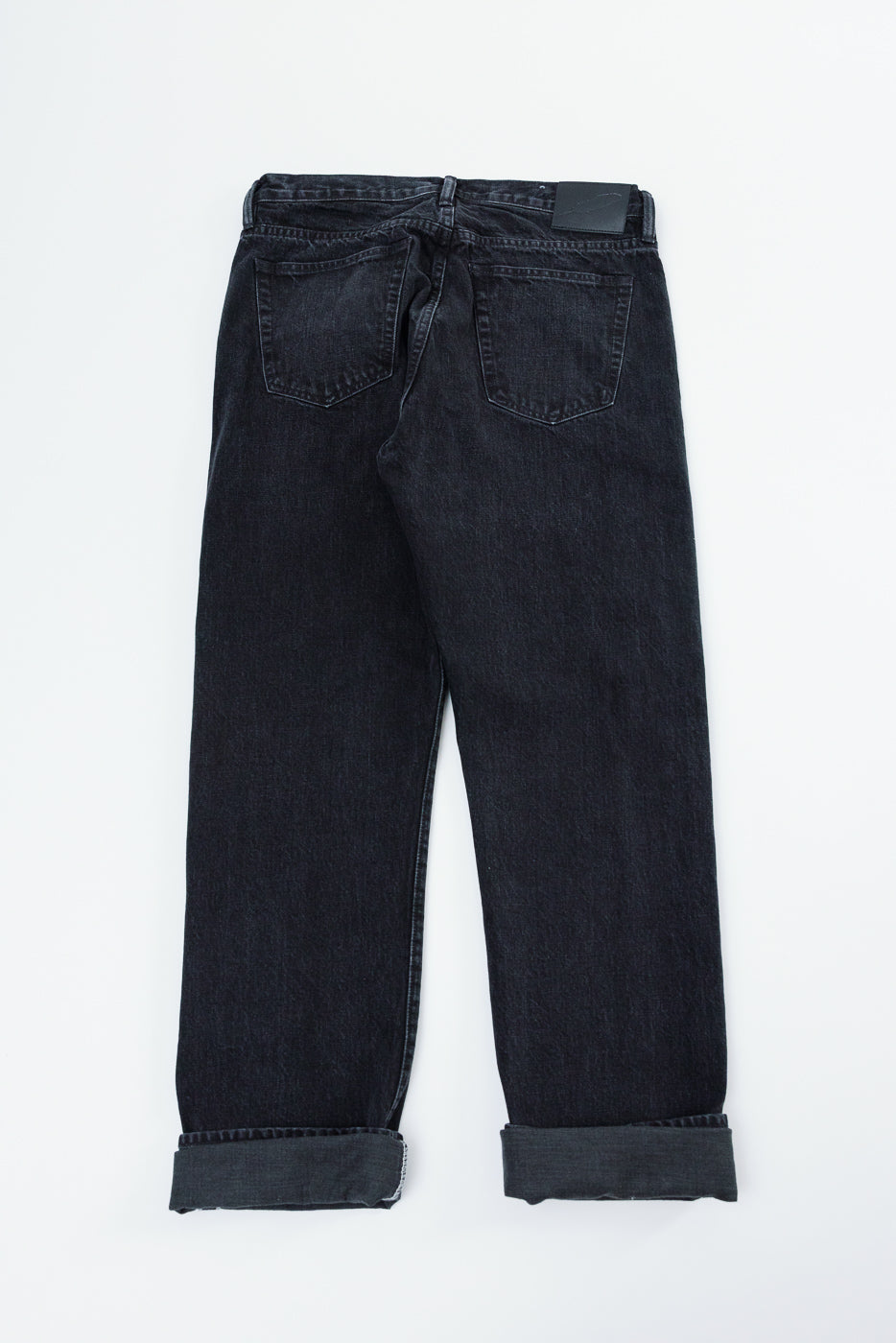 Back view flat lay of dark selvedge denim jeans with cuffed hems, rear pockets, and leather patch