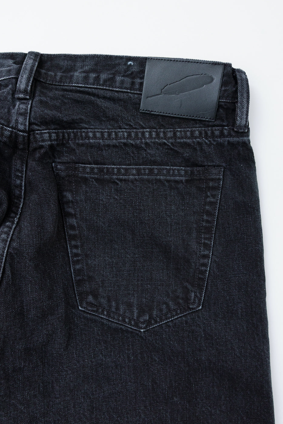 Close-up back view of black selvedge jeans showing rear pocket, stitching, and leather feather patch detail